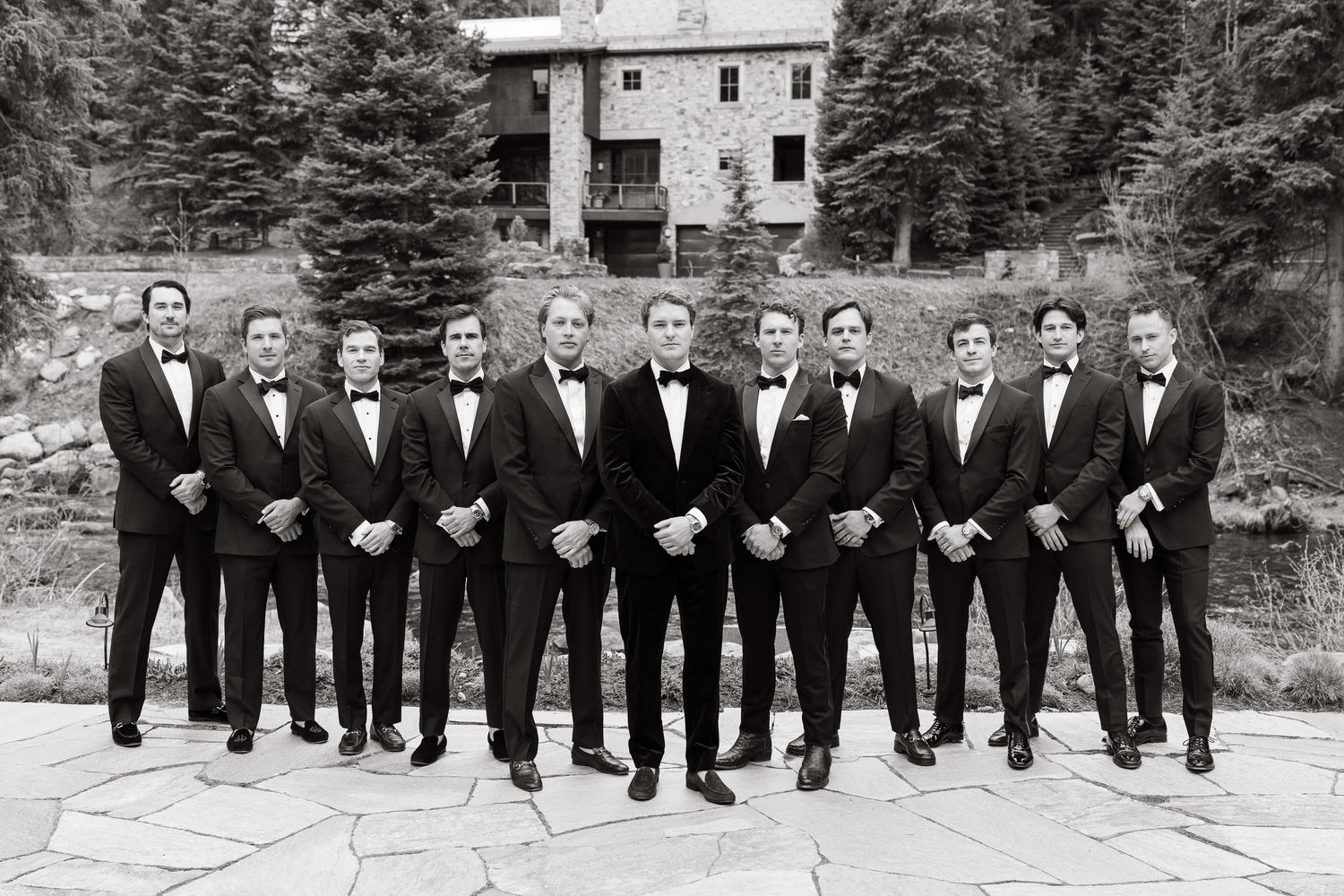 Groom and groomsmen in black tuxedos posing outdoors at a mountain wedding venue in a classic black and white photo.
