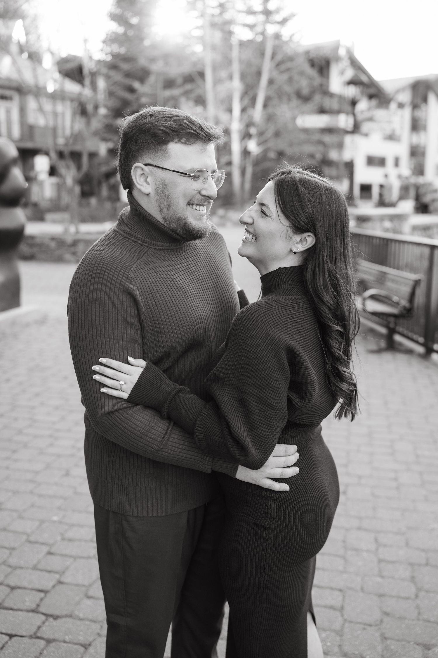 Black and white photo of a laughing couple embracing outdoors in a cozy town square setting.