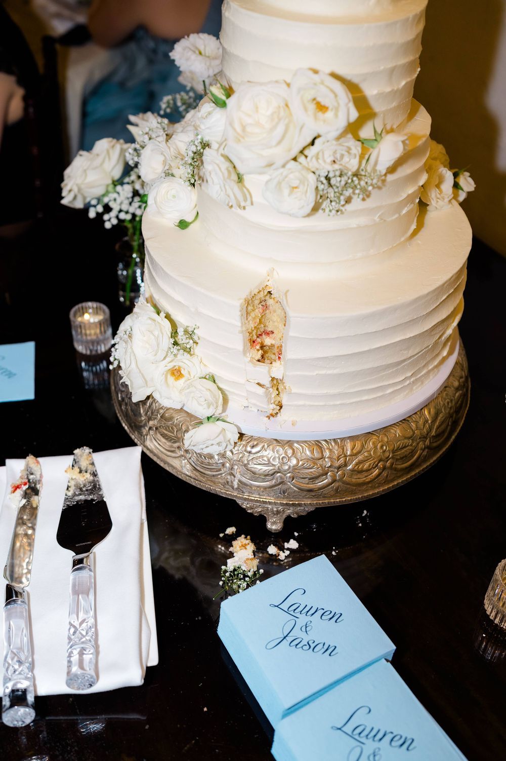 Three-tier white wedding cake with white roses on a glass stand, with a slice cut out, cake server and Lauren & Jason place cards nearby.