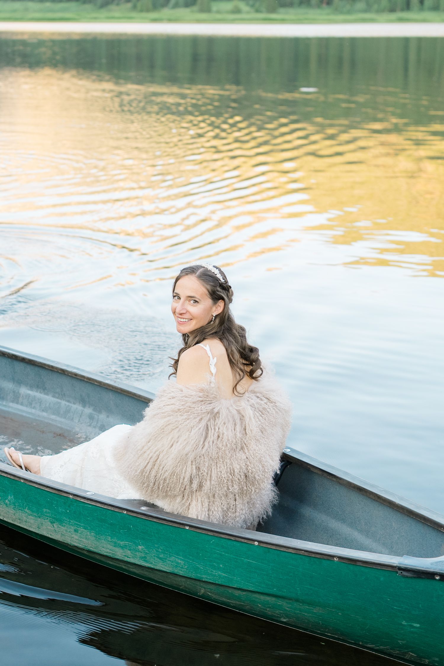 Woman in elegant cream dress reclining in green canoe on tranquil lake during golden hour.