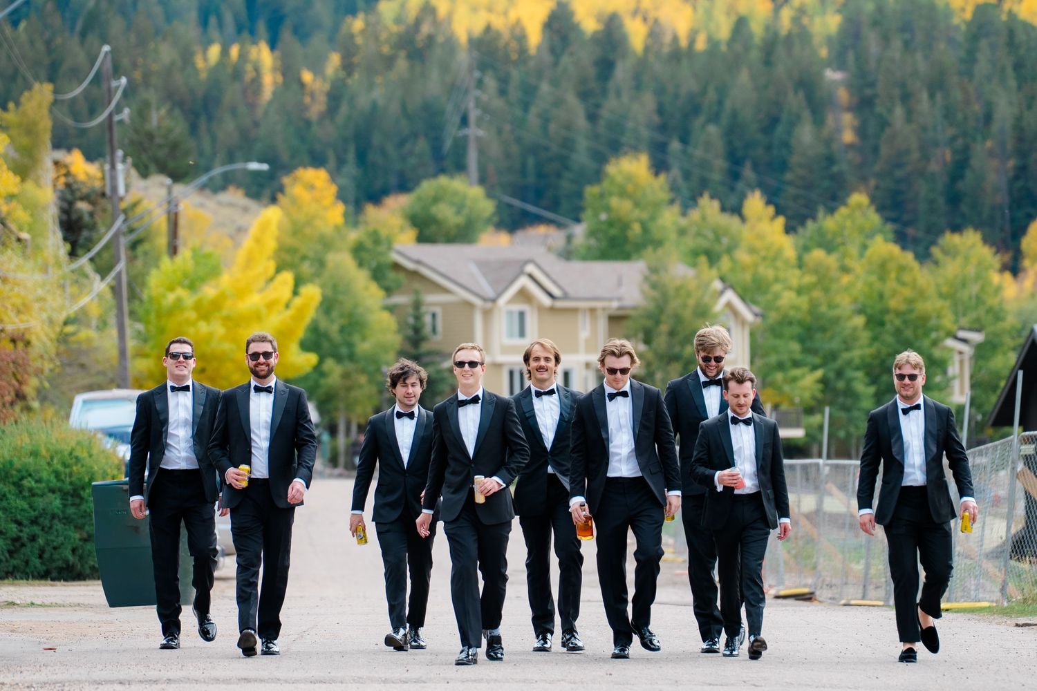 A group in formal black tuxedos walks together on a path with autumn trees and mountains in the background.