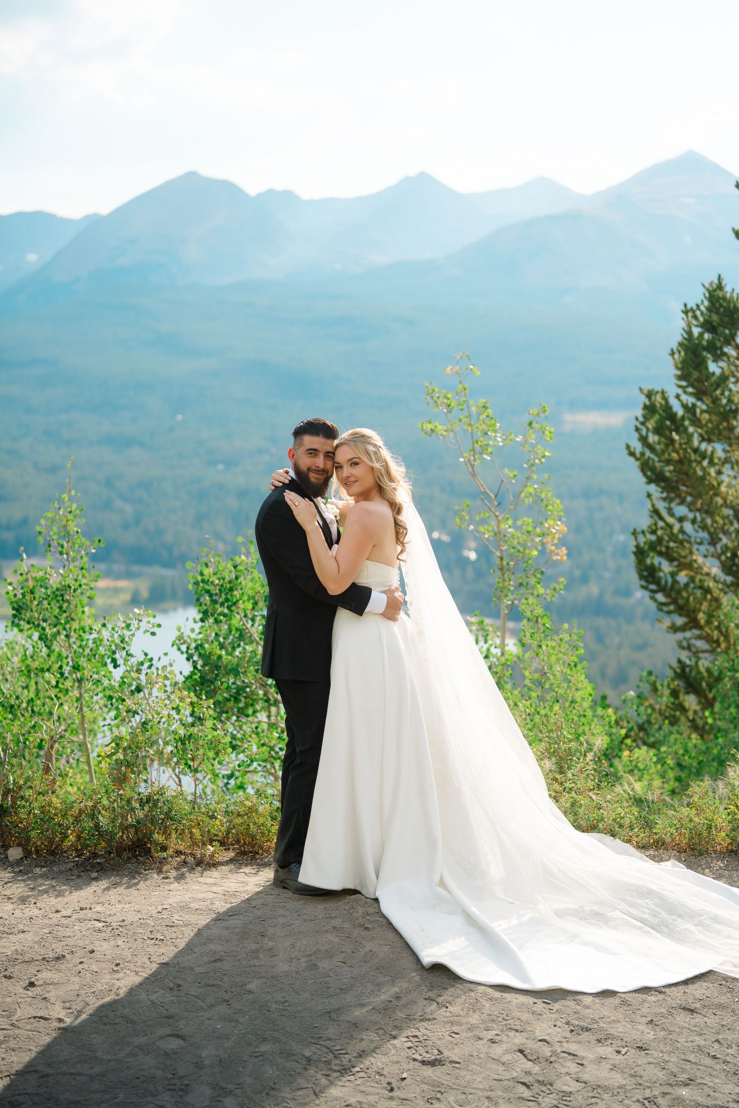 A wedding couple poses together on a scenic mountain overlook with dramatic mountain peaks in the background.
