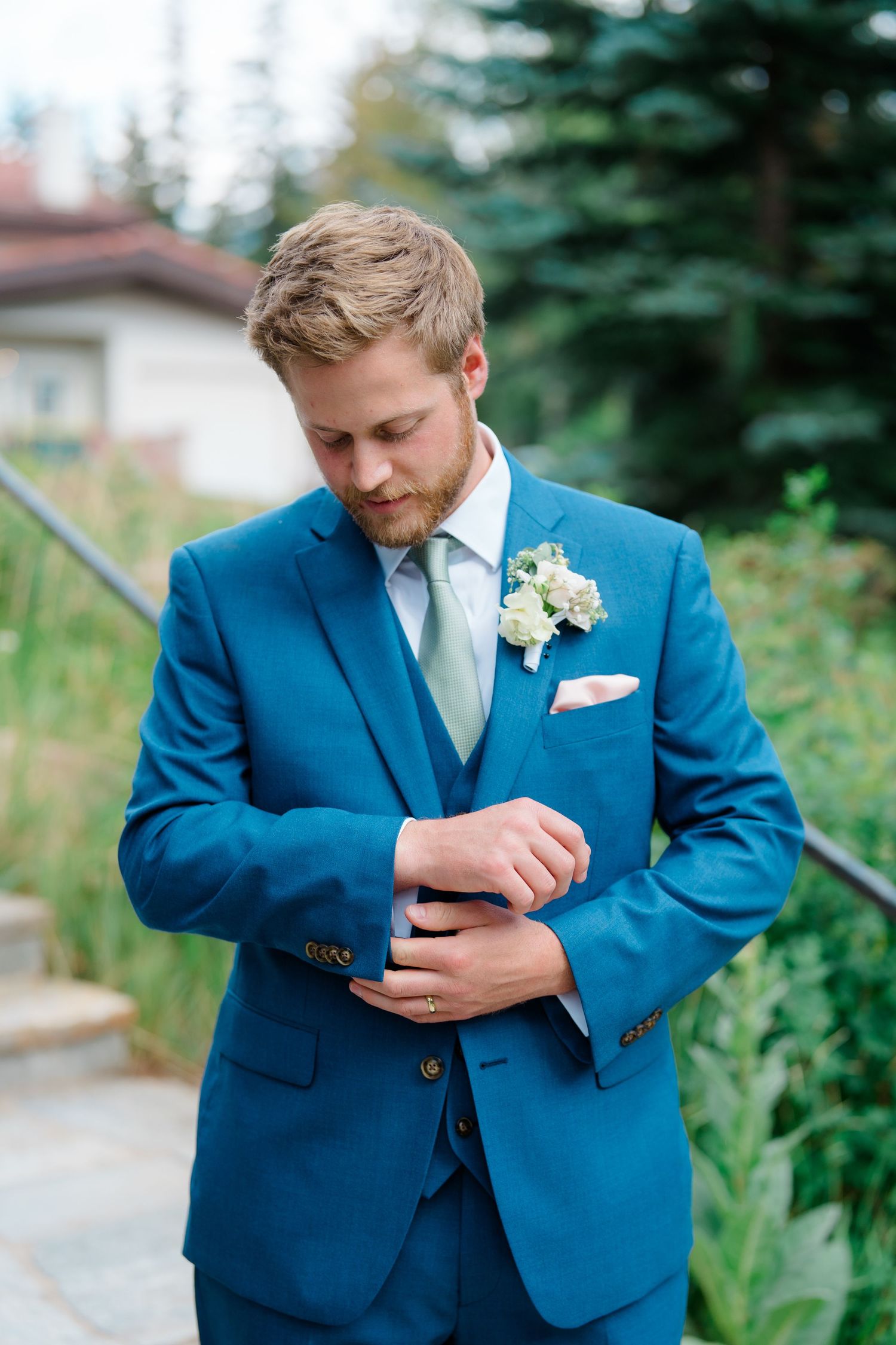 A groom in a bright blue suit adjusts his cuff while standing outdoors with greenery in the background.