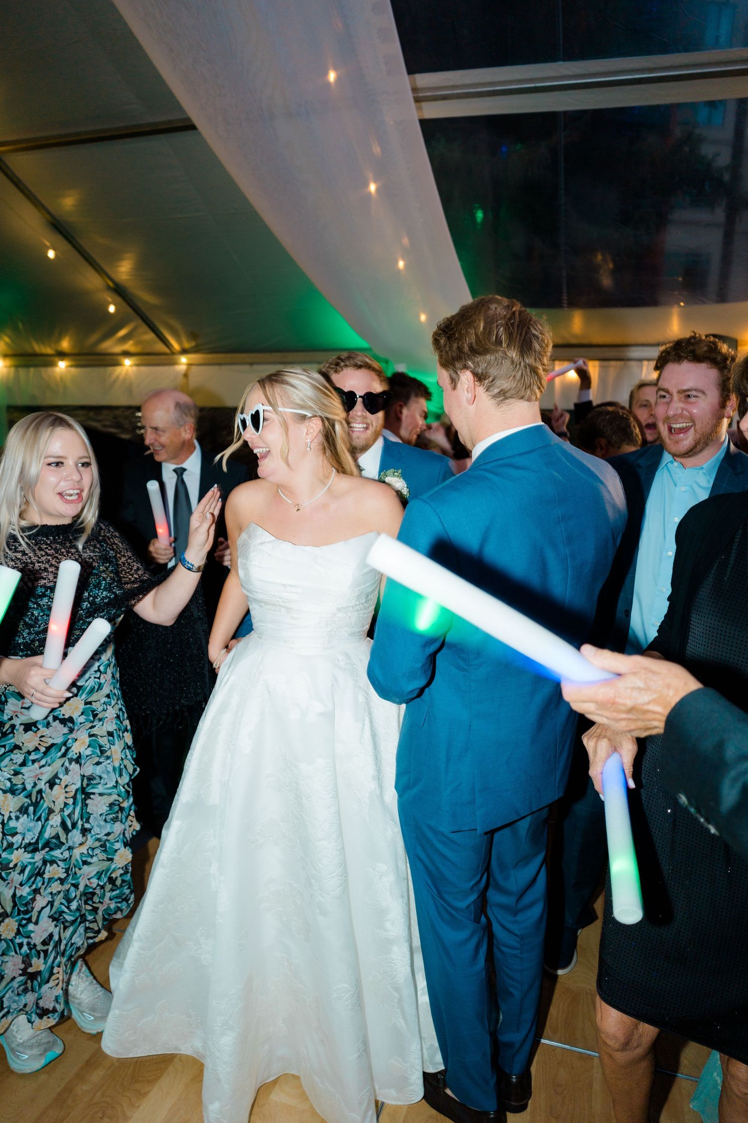 Wedding guests dance and celebrate with glowing foam sticks at a reception under twinkling lights and green uplighting.
