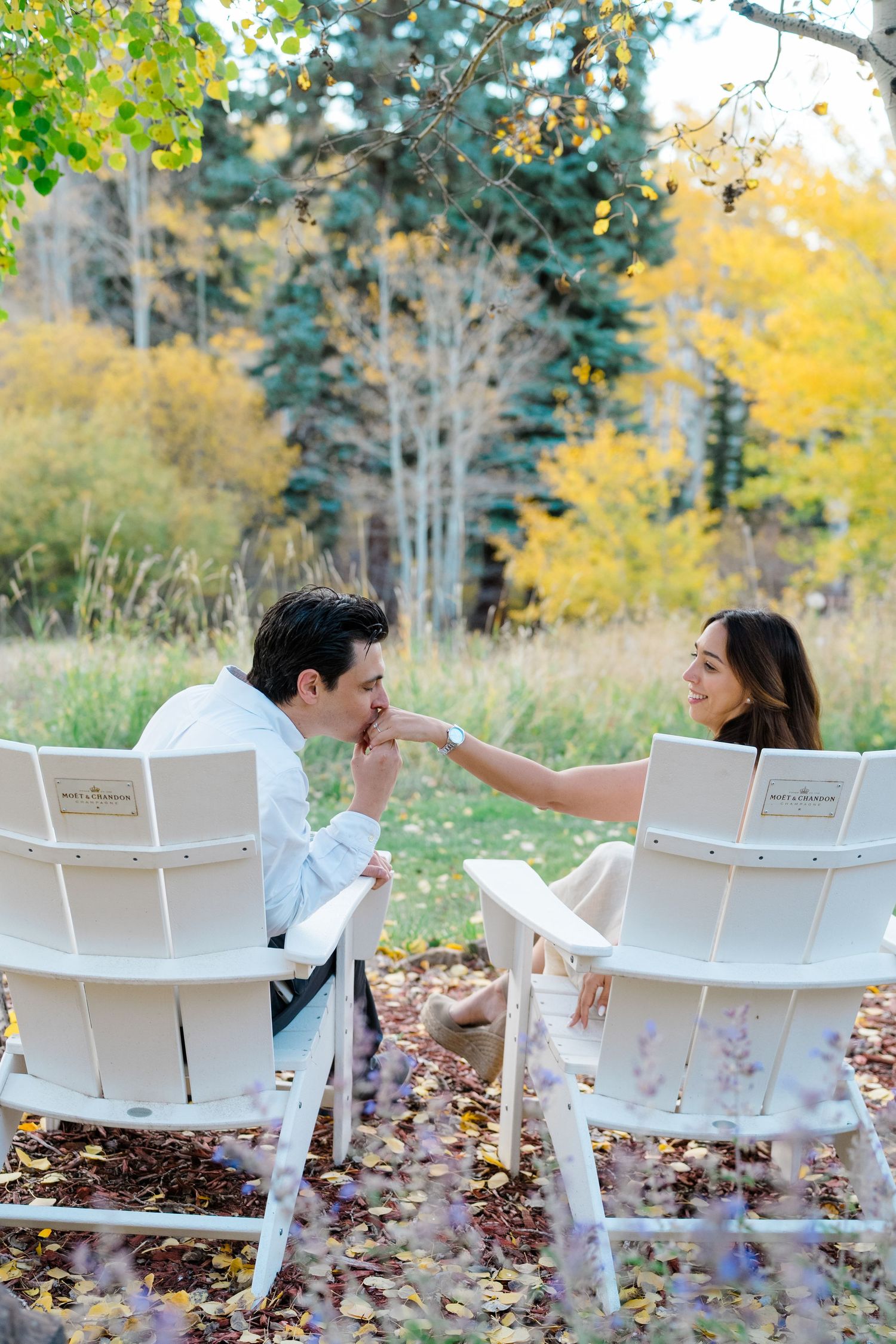 Two white Adirondack chairs sit among fallen autumn leaves as a couple shares a romantic moment in the background.