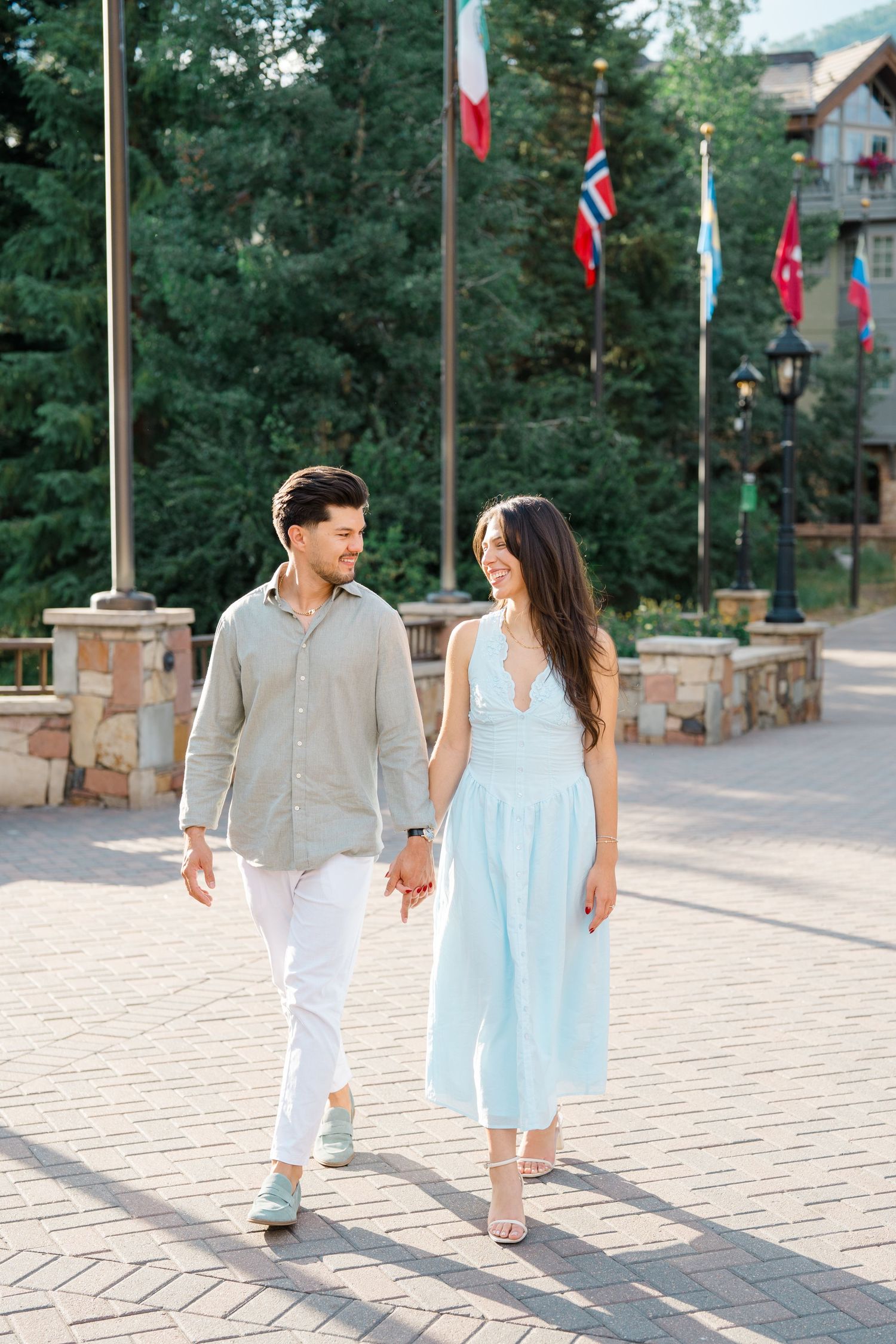 A romantic couple strolls along a stone path with international flags and trees in the background.