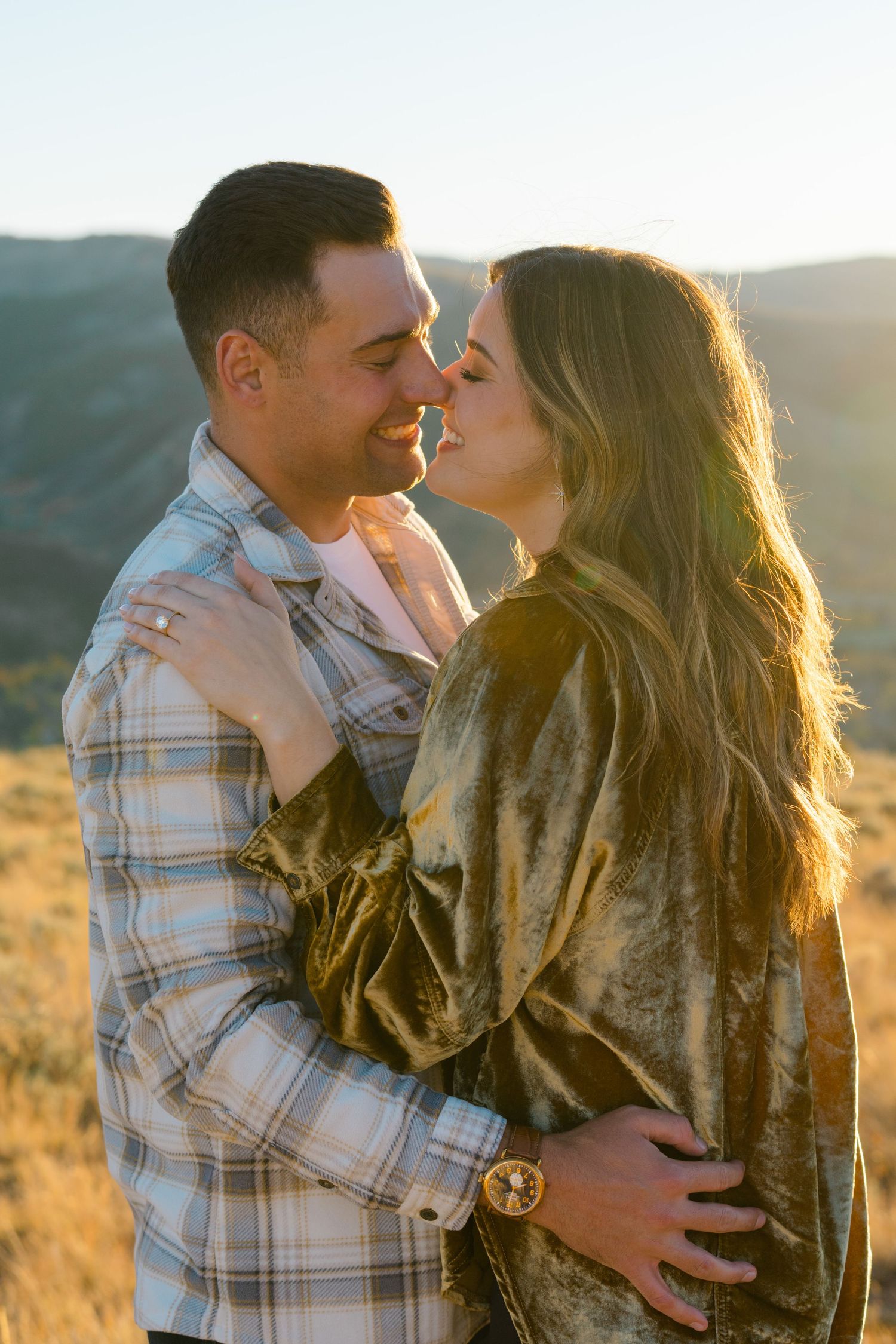 A romantic couple embraces and kisses in golden sunlight during a sunset photo session in nature.