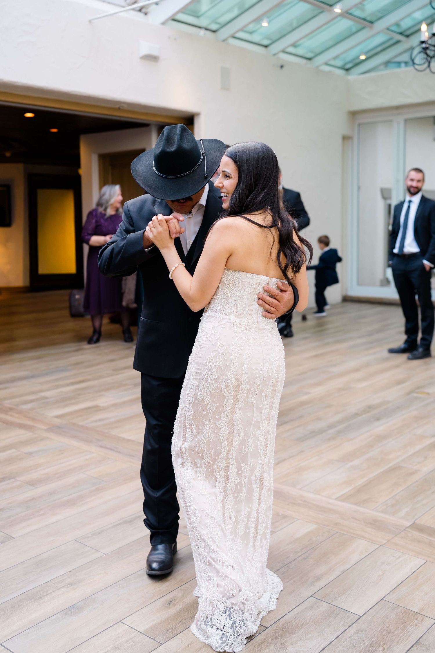 Bride in lace gown dances with groom in black suit and cowboy hat at elegant wedding reception.