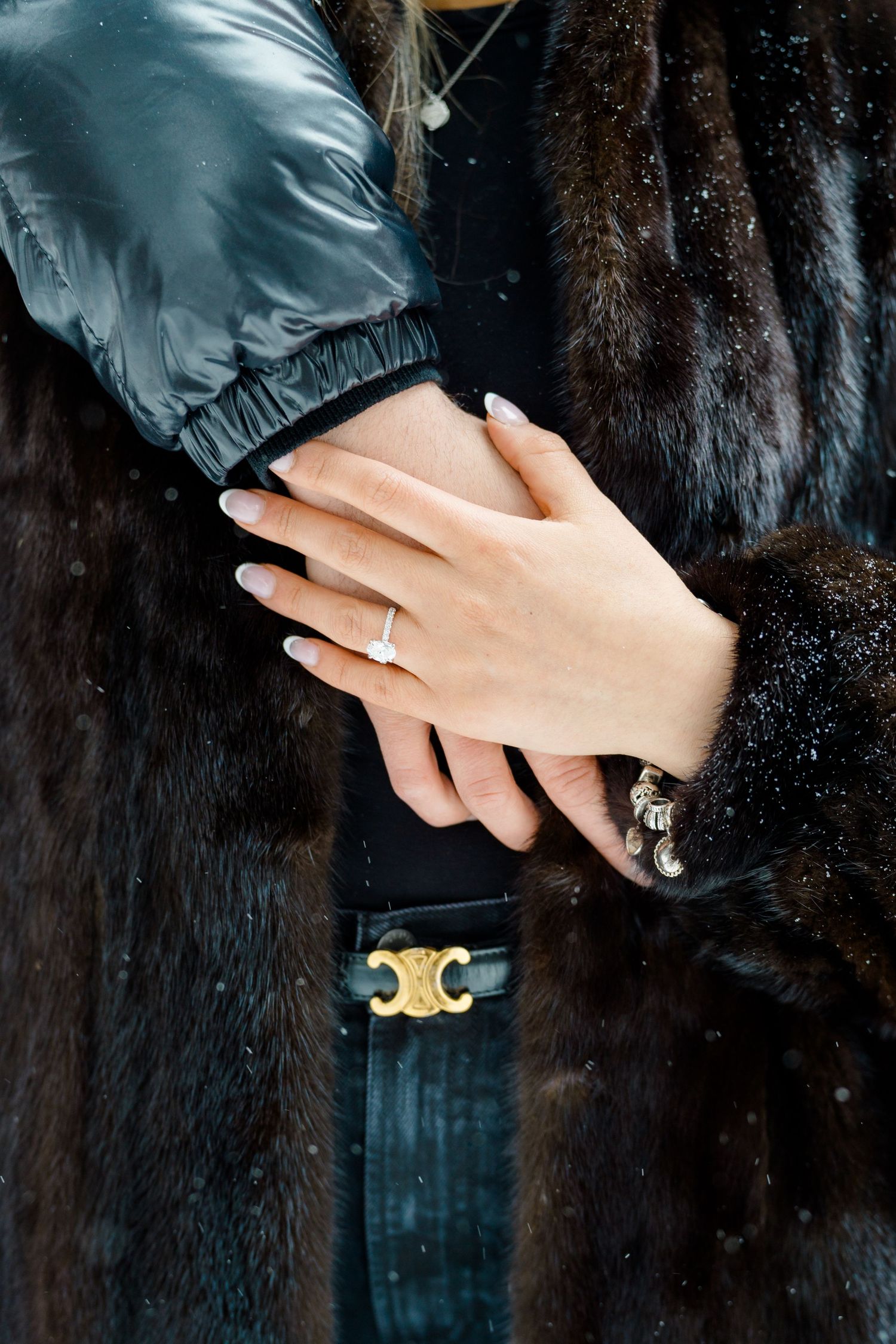 Close-up of a woman's hand wearing a diamond engagement ring, resting on a dark fur coat in snowy weather.
