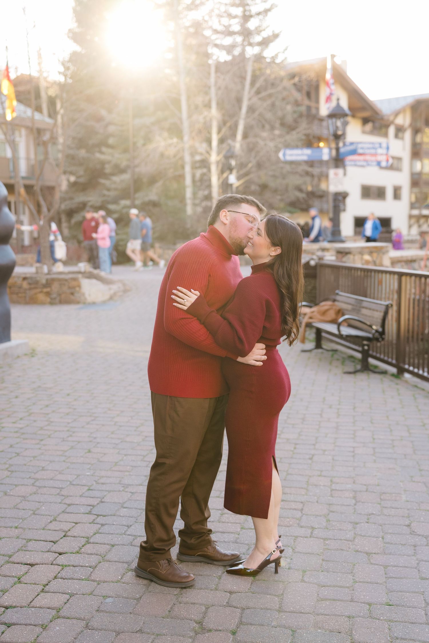 Couple in matching red outfits sharing a kiss at golden hour in a scenic mountain village plaza.