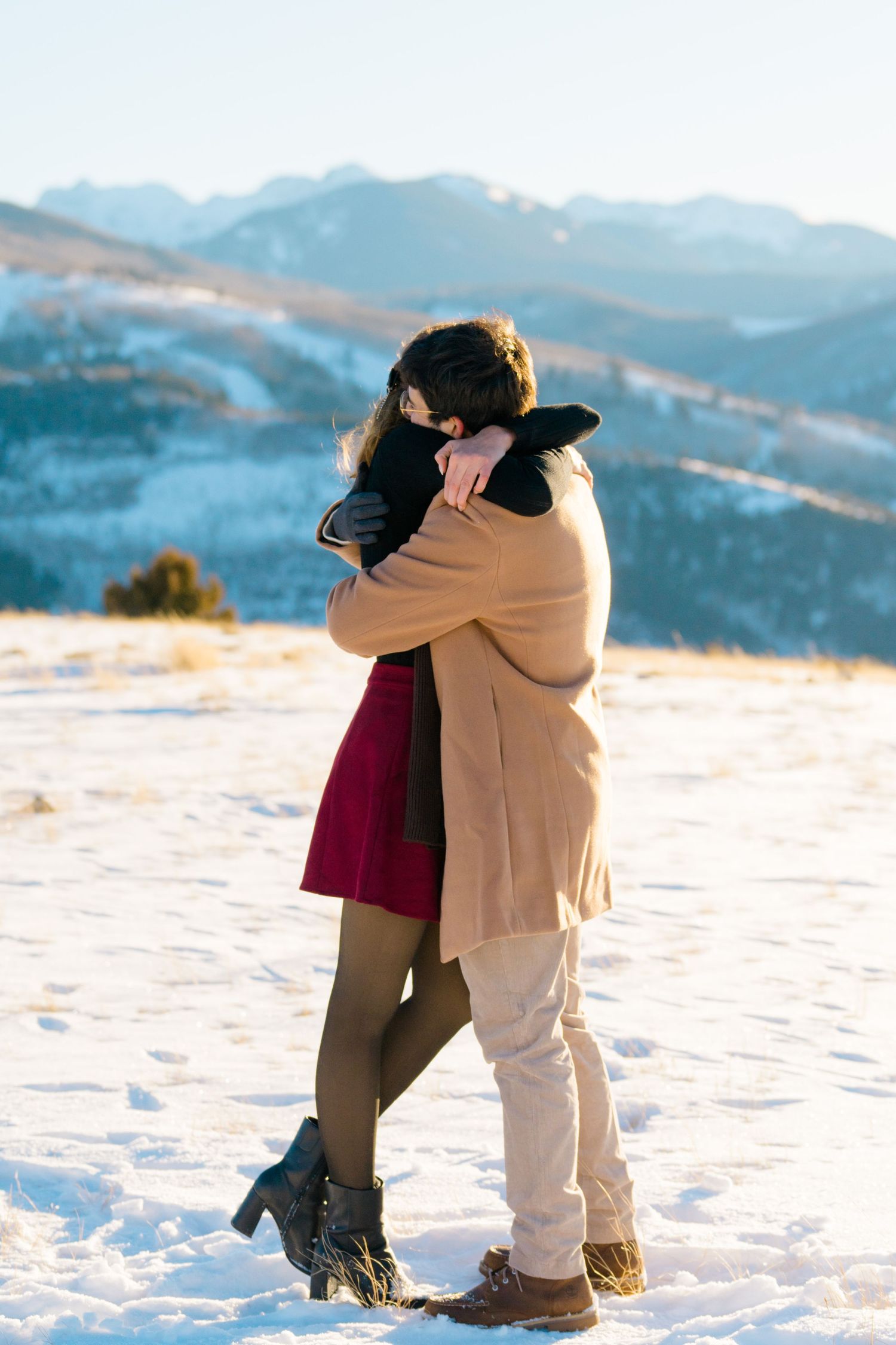 Two people embrace in winter snow with scenic mountain views in the background.