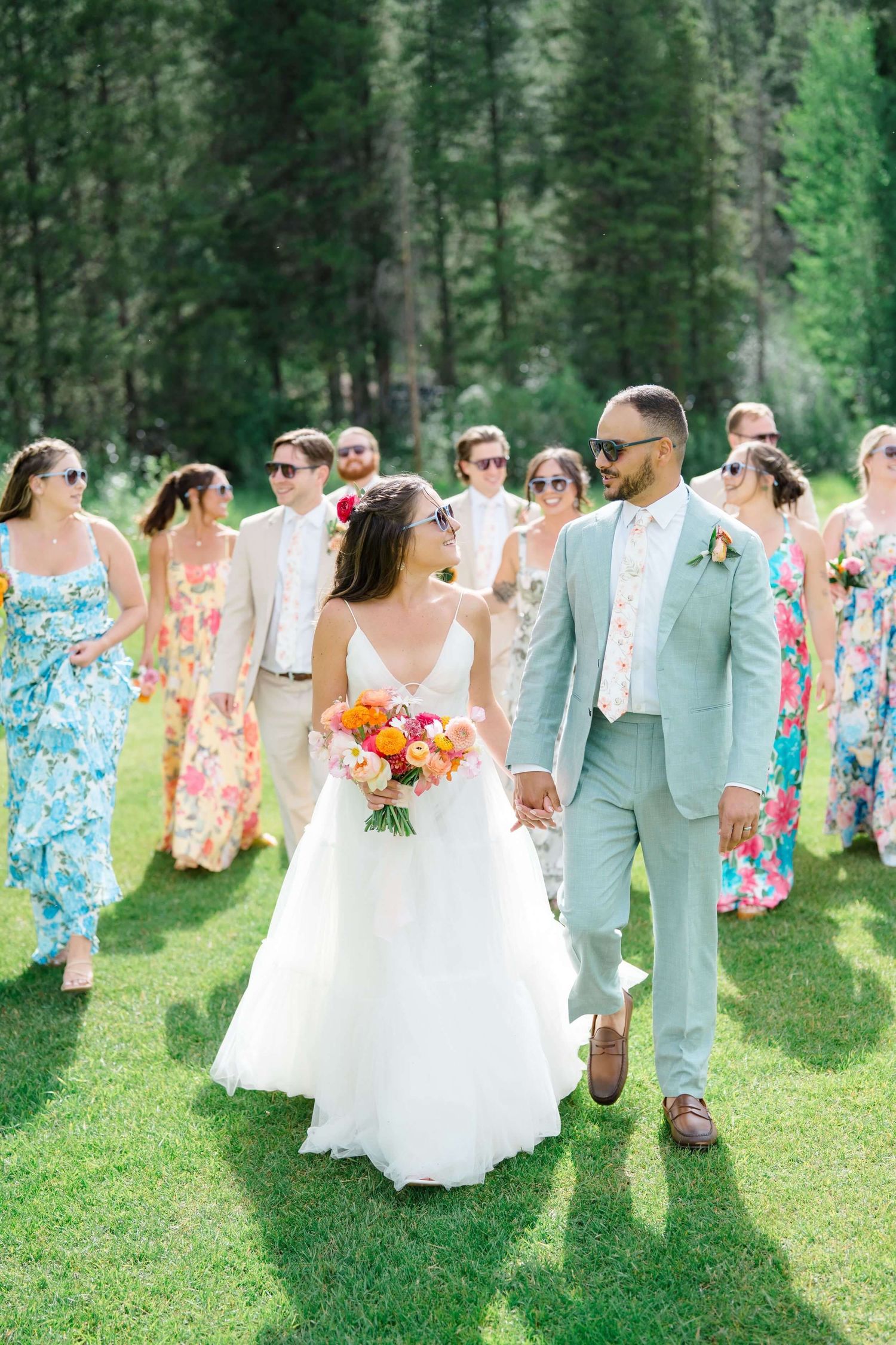 Camp Hale Wedding party in colorful floral dresses walking on grass with mountains and pine trees in background.
