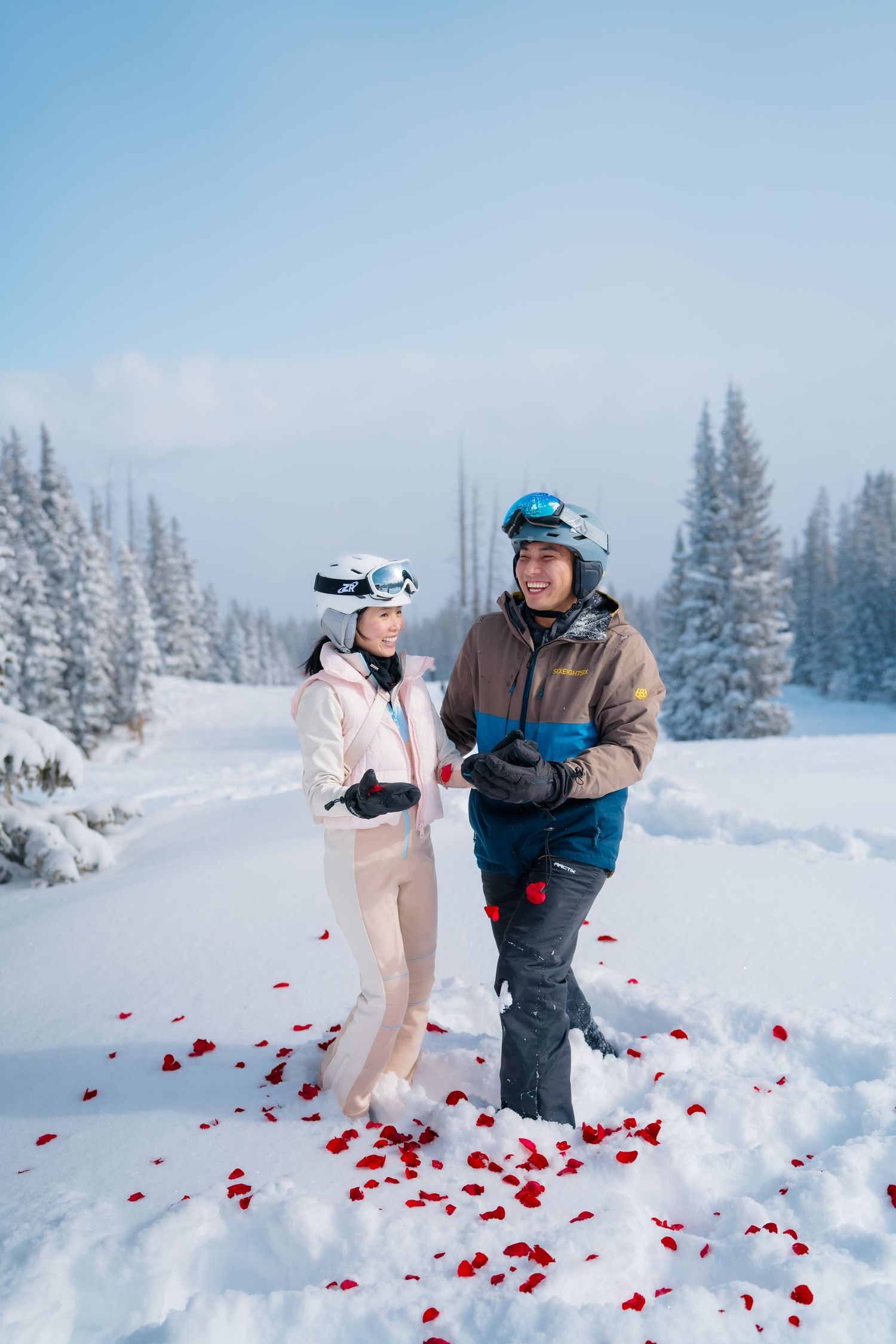 Couple in ski gear standing in snowy mountain landscape with red rose petals scattered around them in winter.