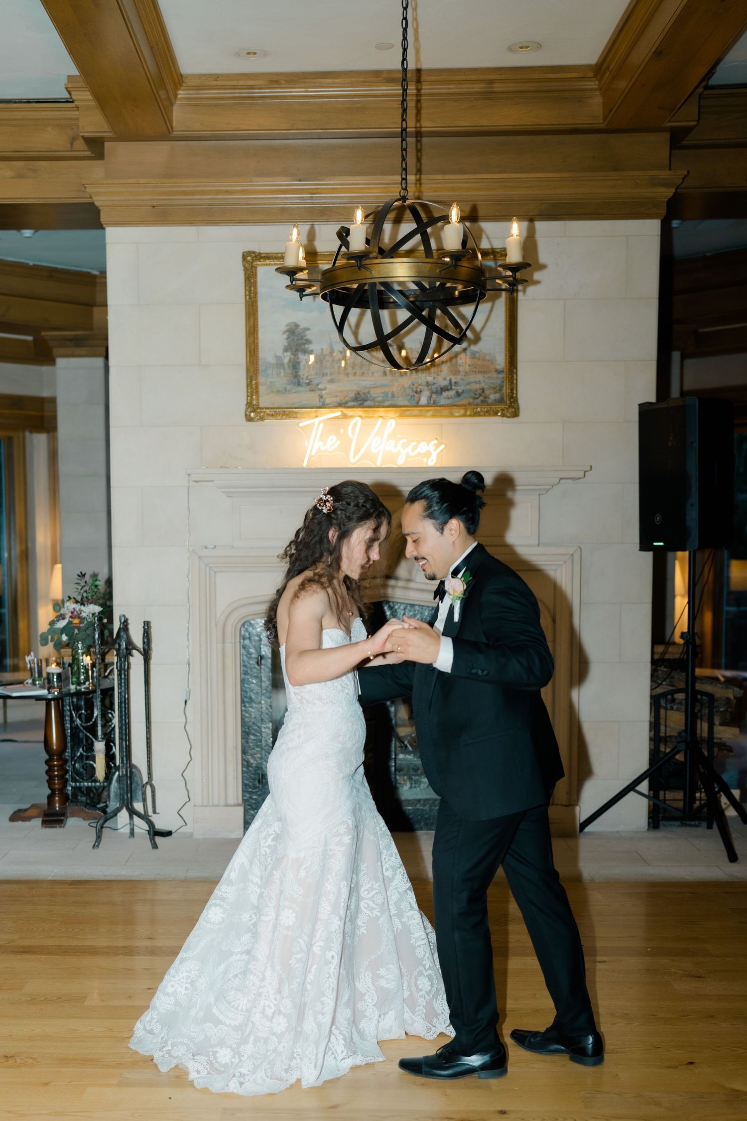 A couple dances together in a spacious ballroom with wooden floors and elegant chandeliers overhead.