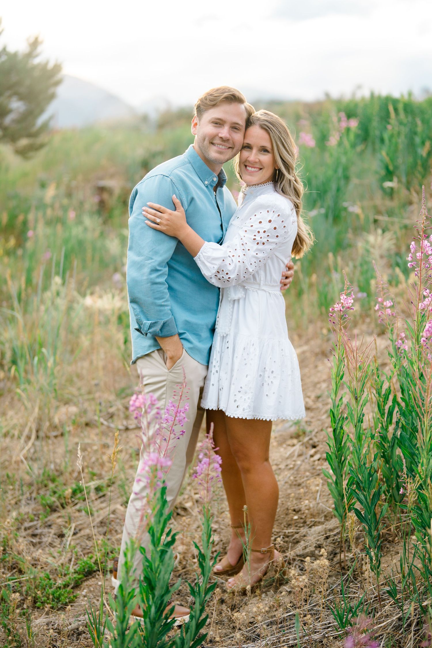 A couple embraces among wildflowers with mountains visible in the distance.