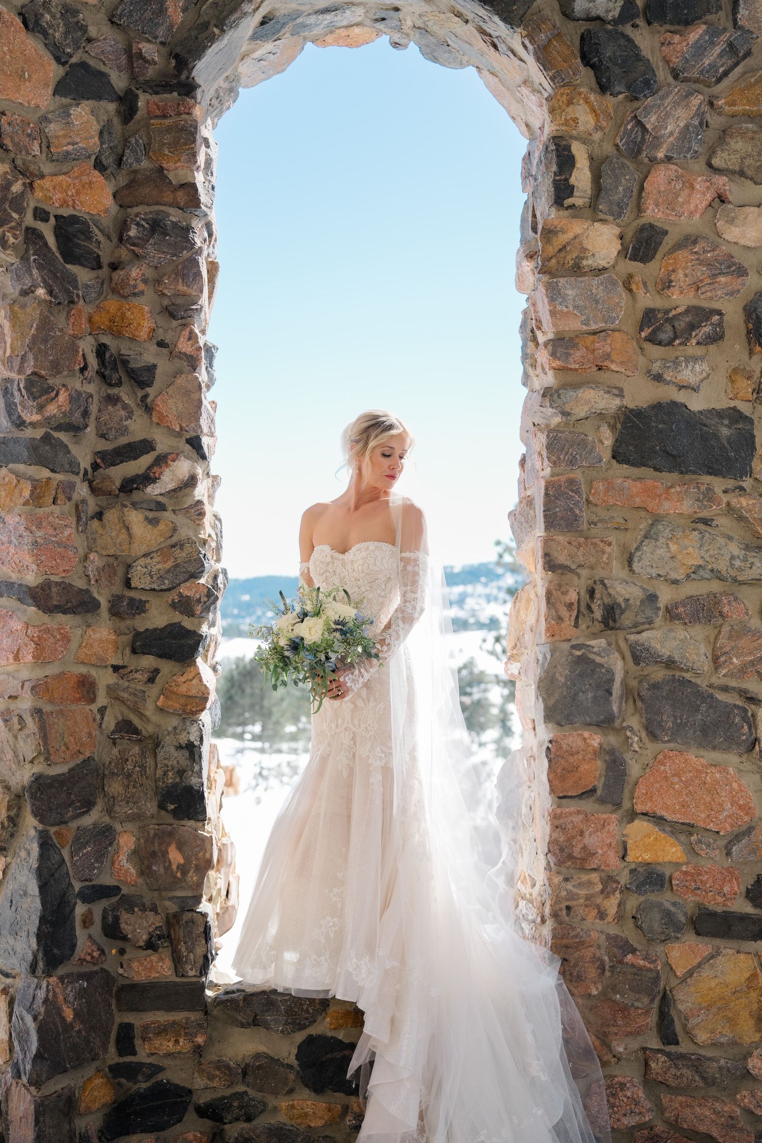 A bride in a flowing white gown poses in stone archways overlooking a scenic mountain landscape.
