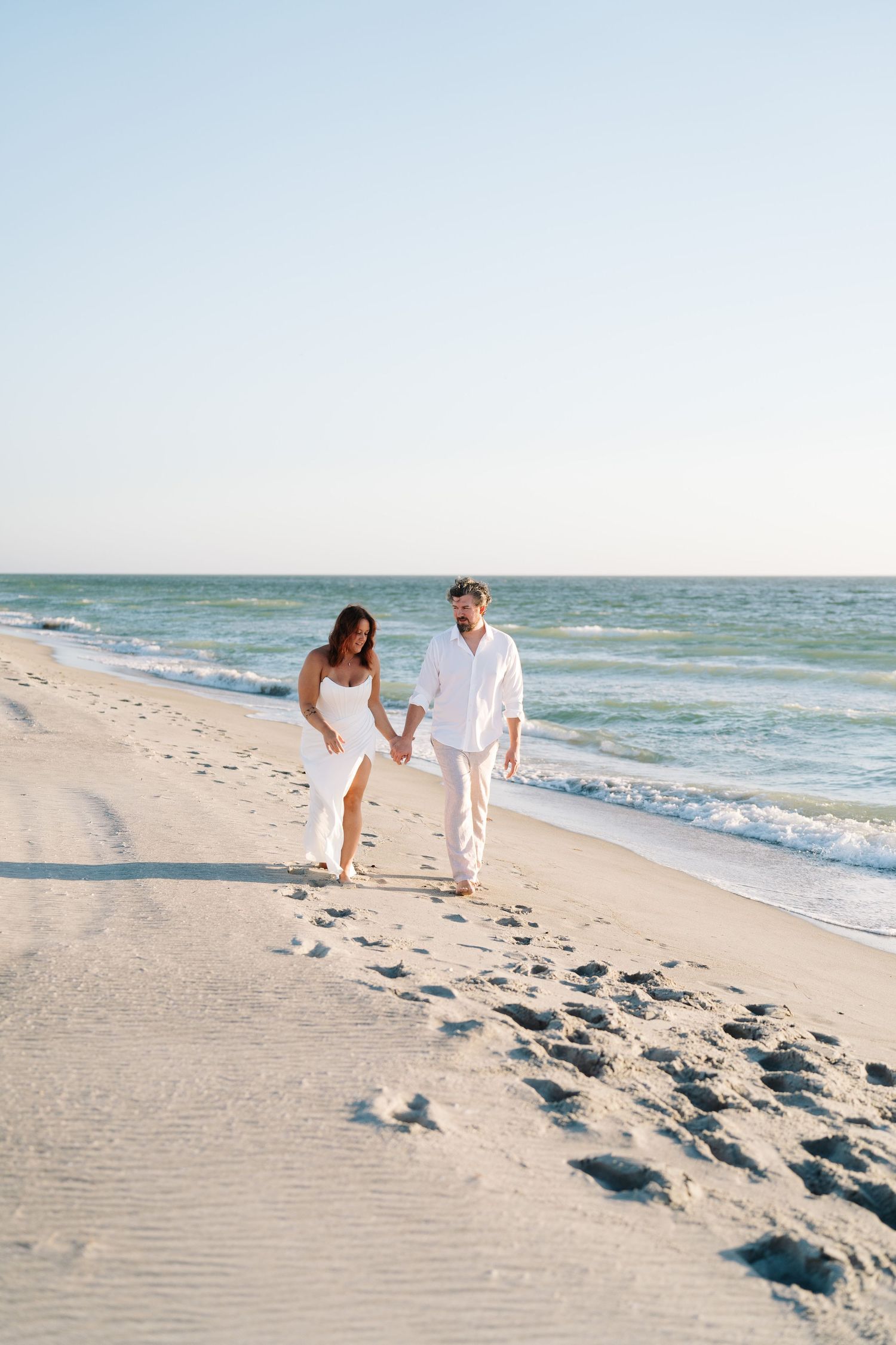 A couple in white outfits walking together along a sunny beach shoreline with waves in the background.
