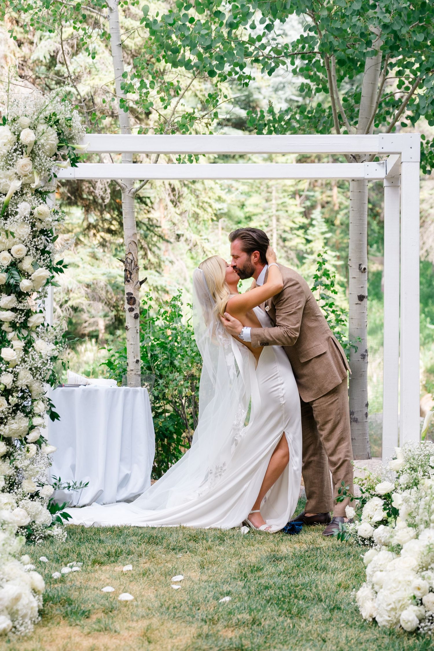 Bride and groom share first kiss under white floral arch at outdoor garden wedding ceremony.