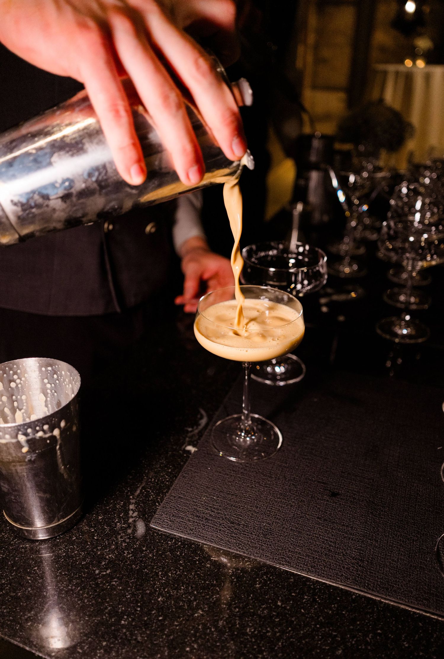 Bartender pouring creamy espresso martini cocktail from shaker into coupe glass at dark bar.