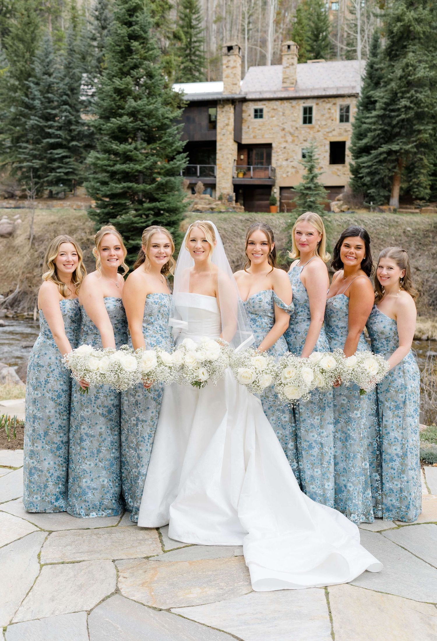 Bride in white gown with seven bridesmaids in blue floral dresses holding white bouquets outdoors near a stone building.