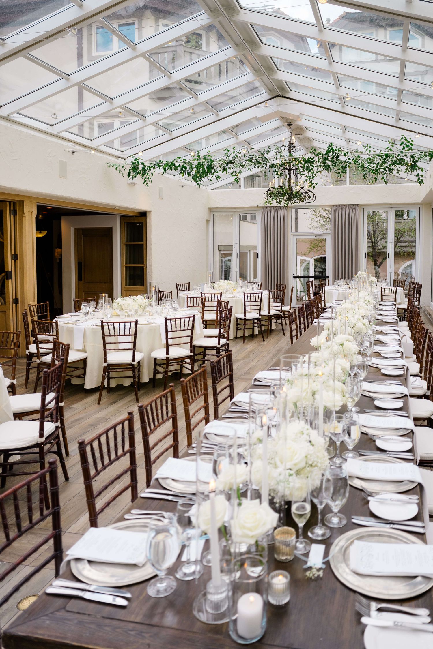 Elegant wedding reception in a glass-ceiling venue with long wooden tables, white floral centerpieces, and chiavari chairs.