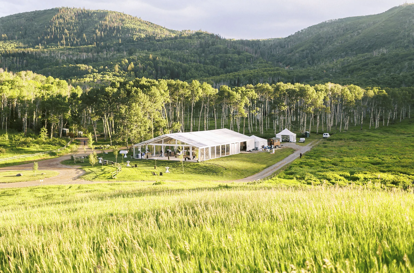 White event tent nestled in lush green meadow surrounded by aspen trees and mountain landscape on a sunny day.