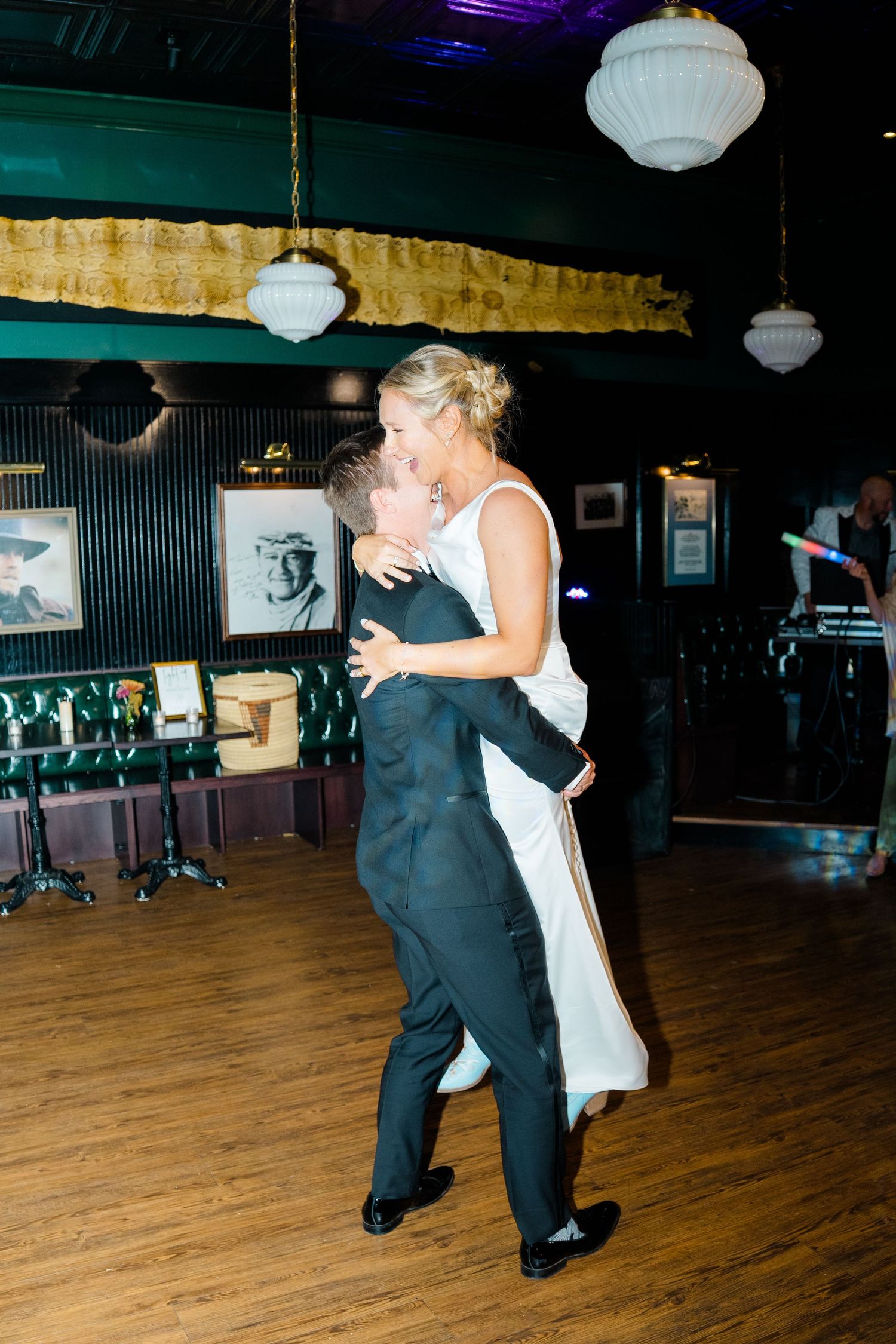 Wedding couple dances together on wooden floor under white paper lanterns in dimly lit venue.