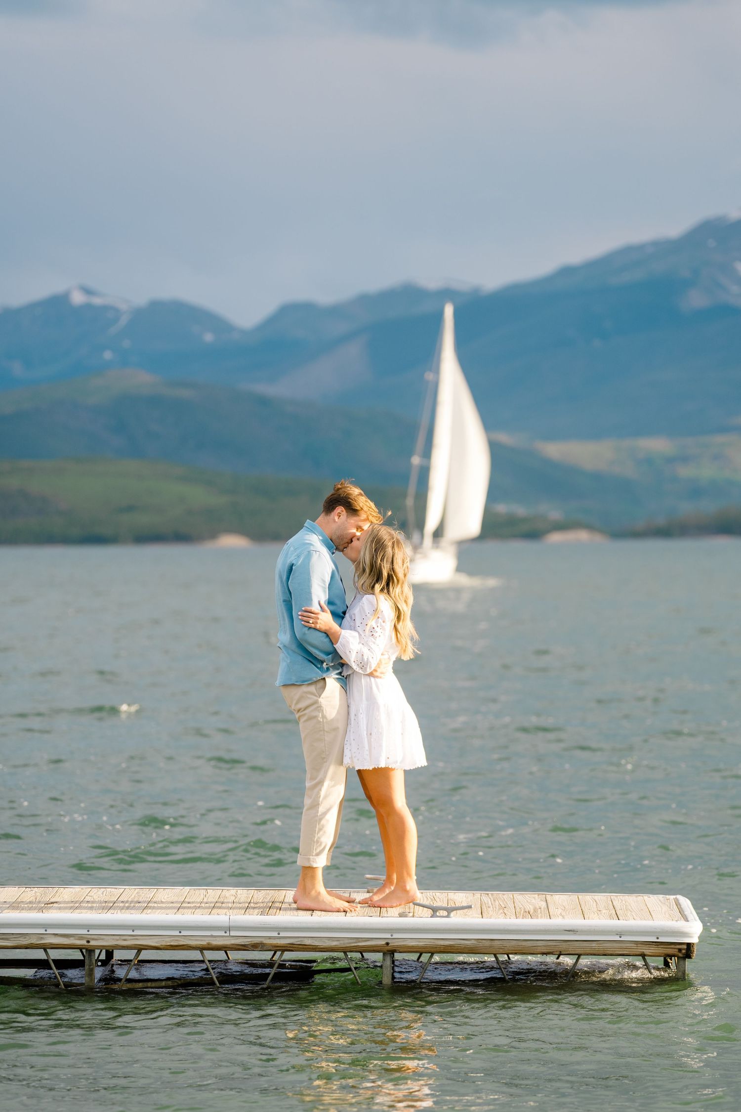 A couple embraces on a wooden dock overlooking a lake with mountains and sailboats in the background.