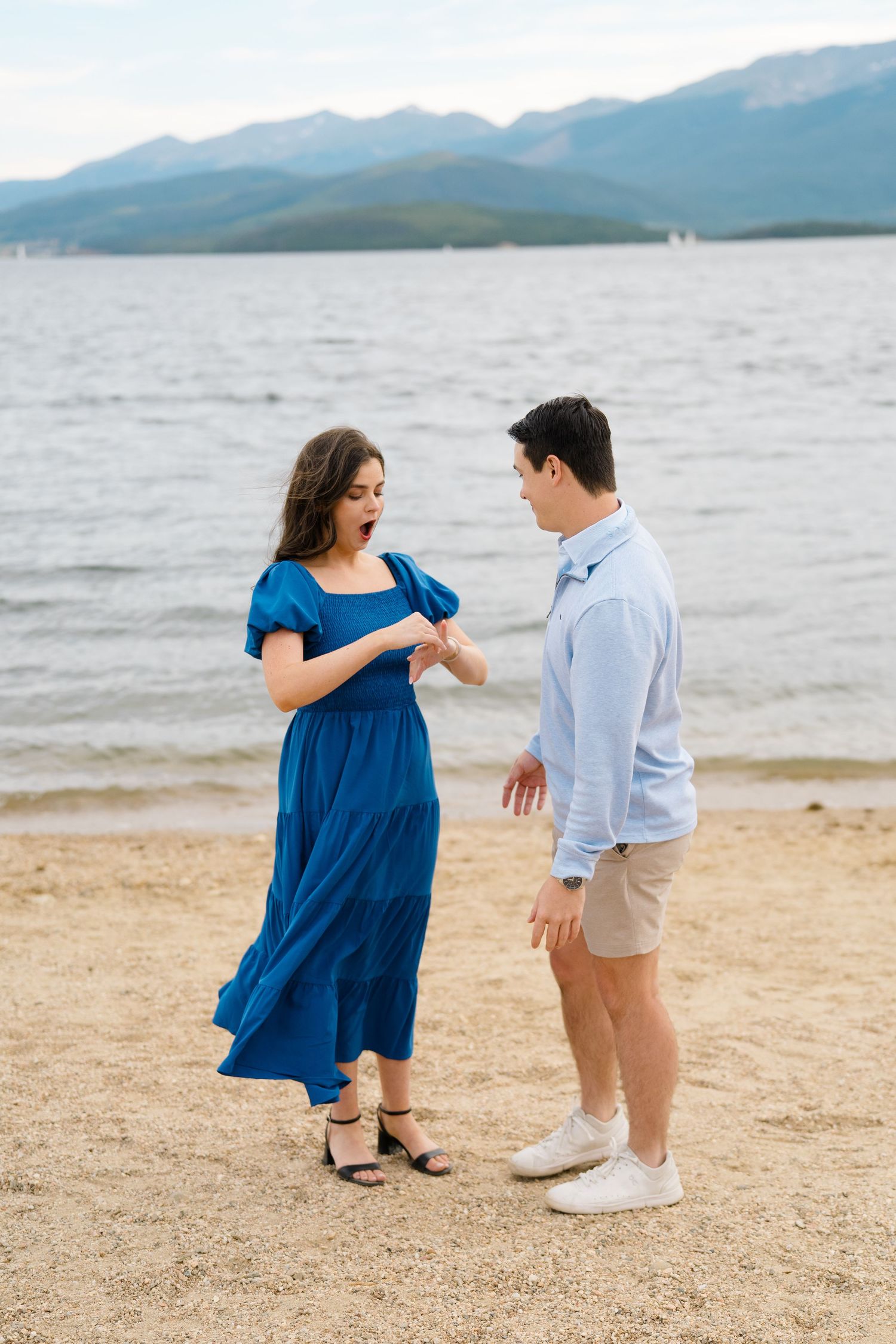 A couple shares a playful moment on a sandy beach with mountains and water in the distance.