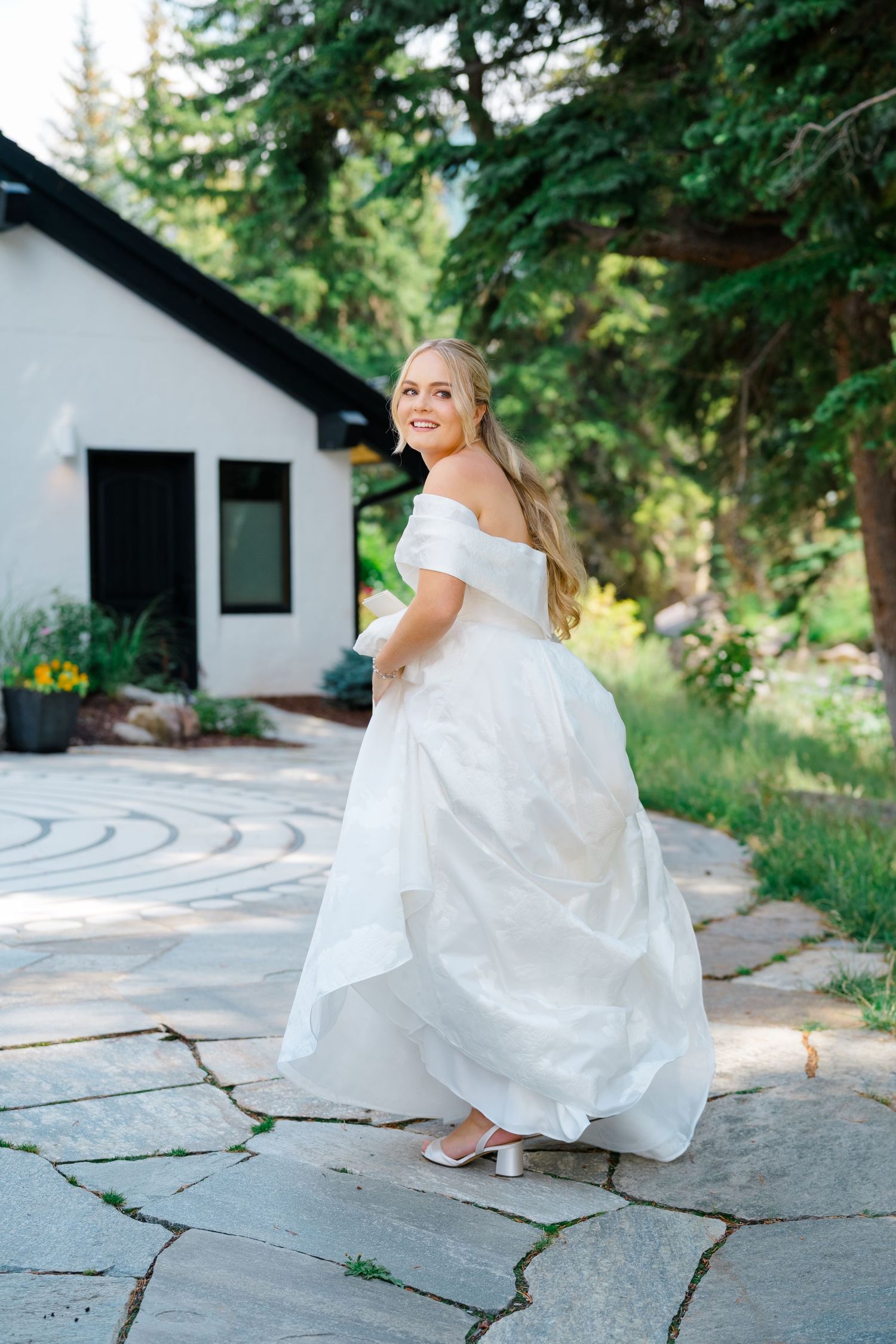 A bride in a flowing white wedding dress twirls and poses on a stone patio surrounded by evergreen trees.