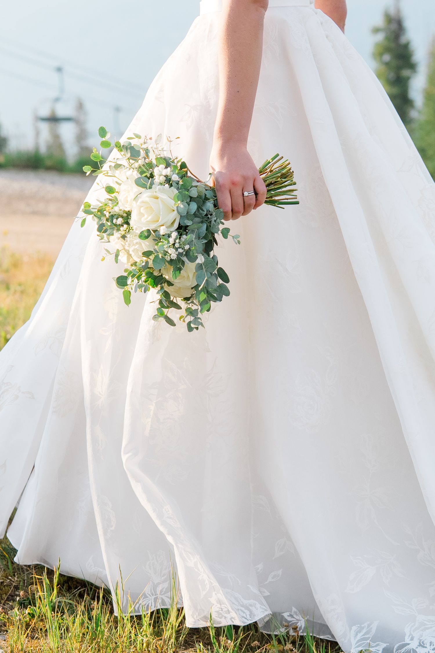 A flowing white wedding dress skirt sways in a grassy field with delicate bouquet details.