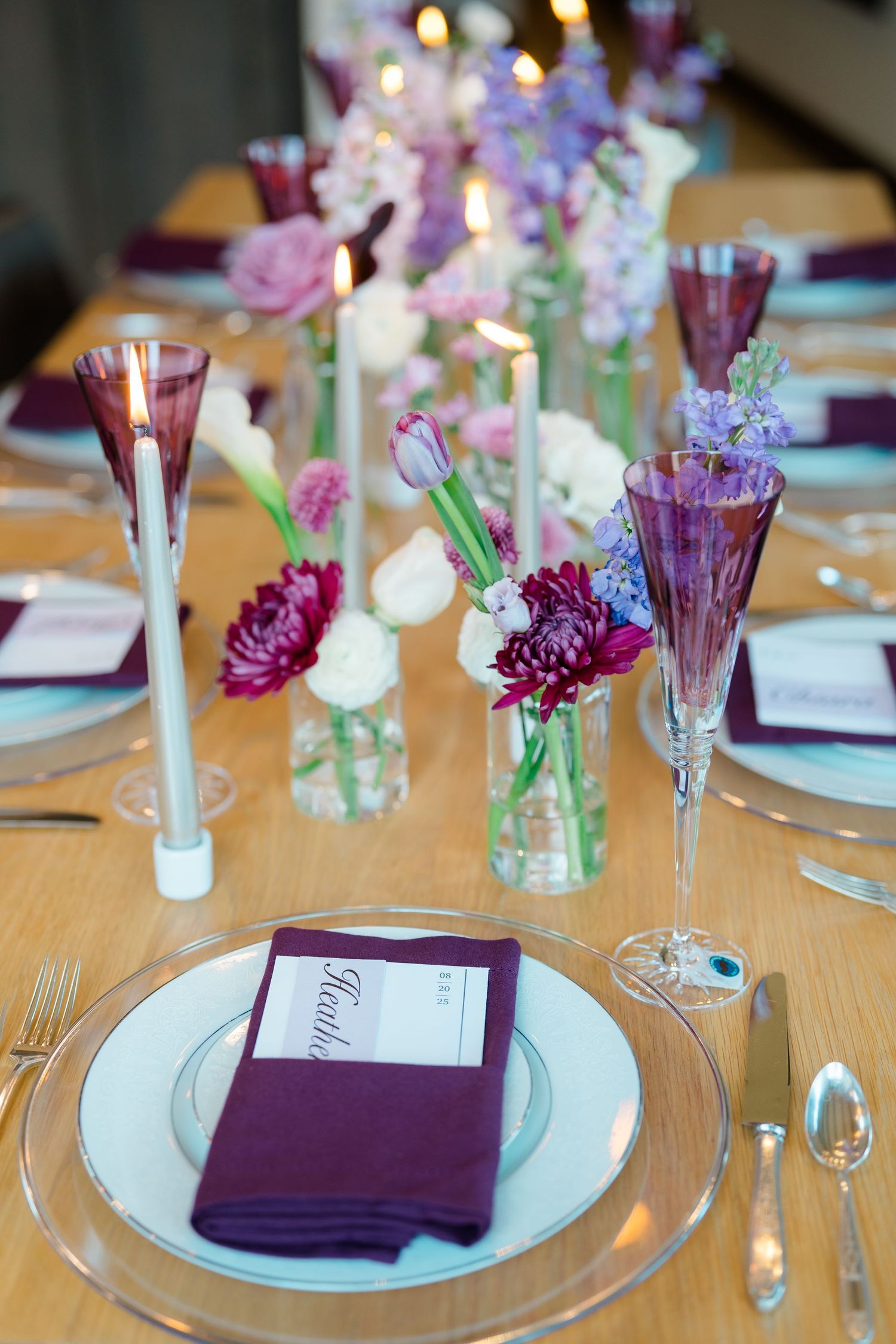 Elegant table setting with purple napkins, clear glass plates, and small floral centerpieces in glass vases.