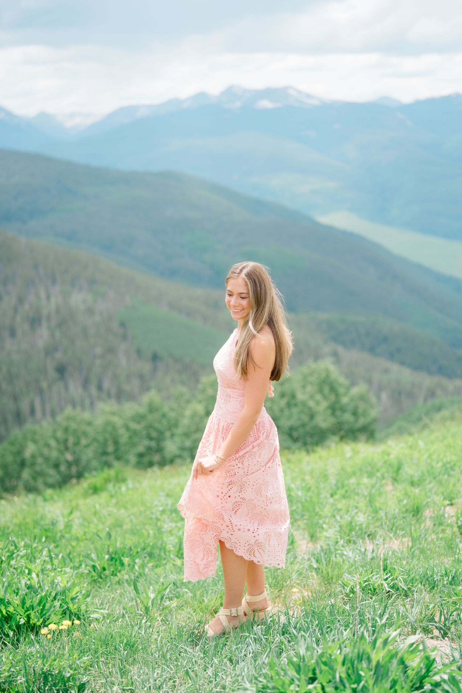 A pink lace dress flows in the mountain breeze against a backdrop of majestic peaks and green valleys during vail senior photography session