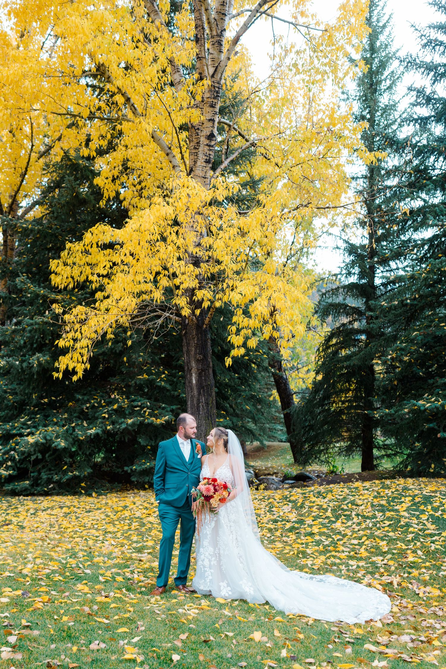 Bride and groom stand among golden fall foliage, her lace gown trailing on leaf-covered grass beneath a vibrant yellow tree.