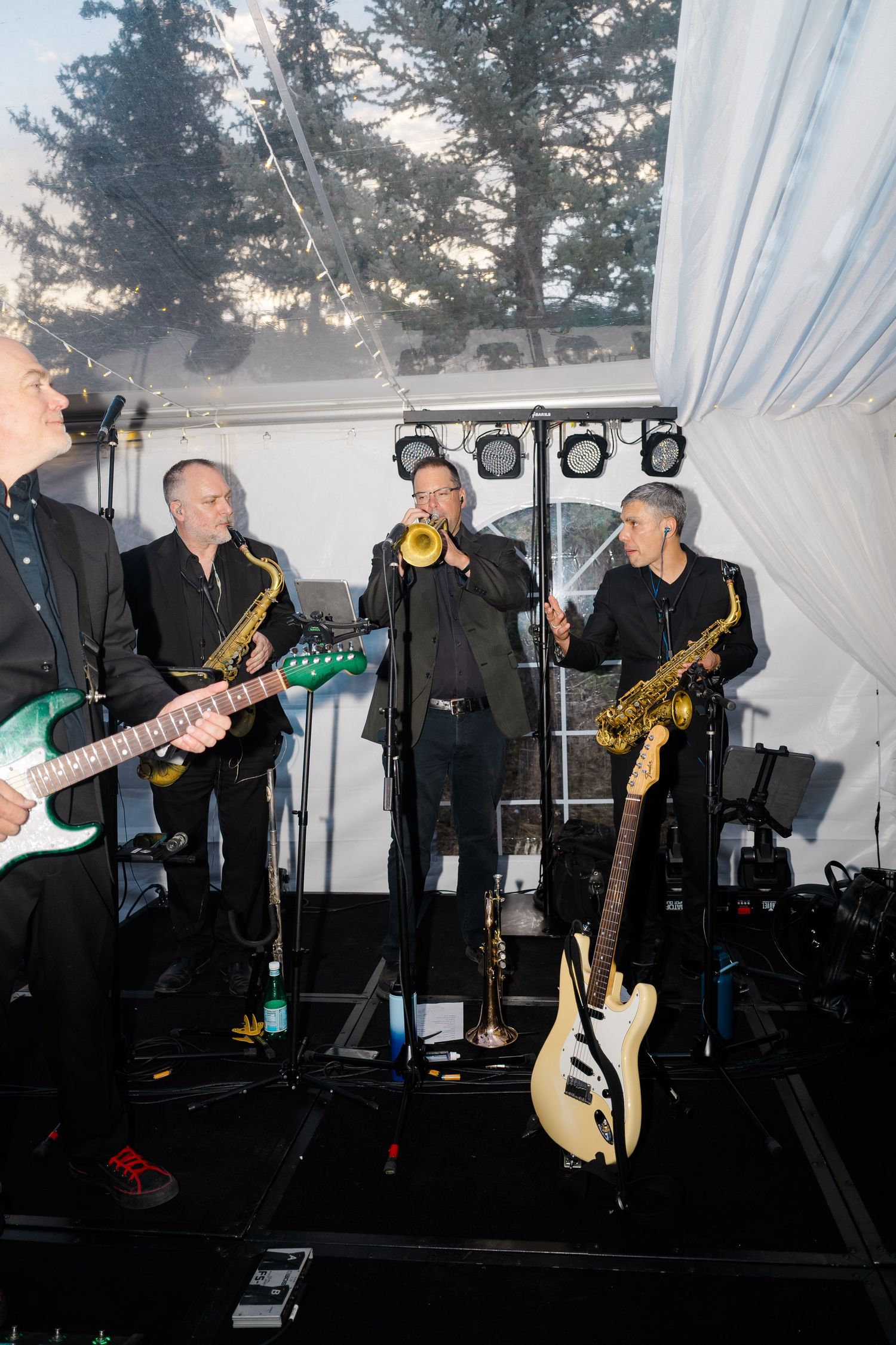 A live band performs in a tent venue with guitarists and saxophonists playing on stage with stage lighting overhead.