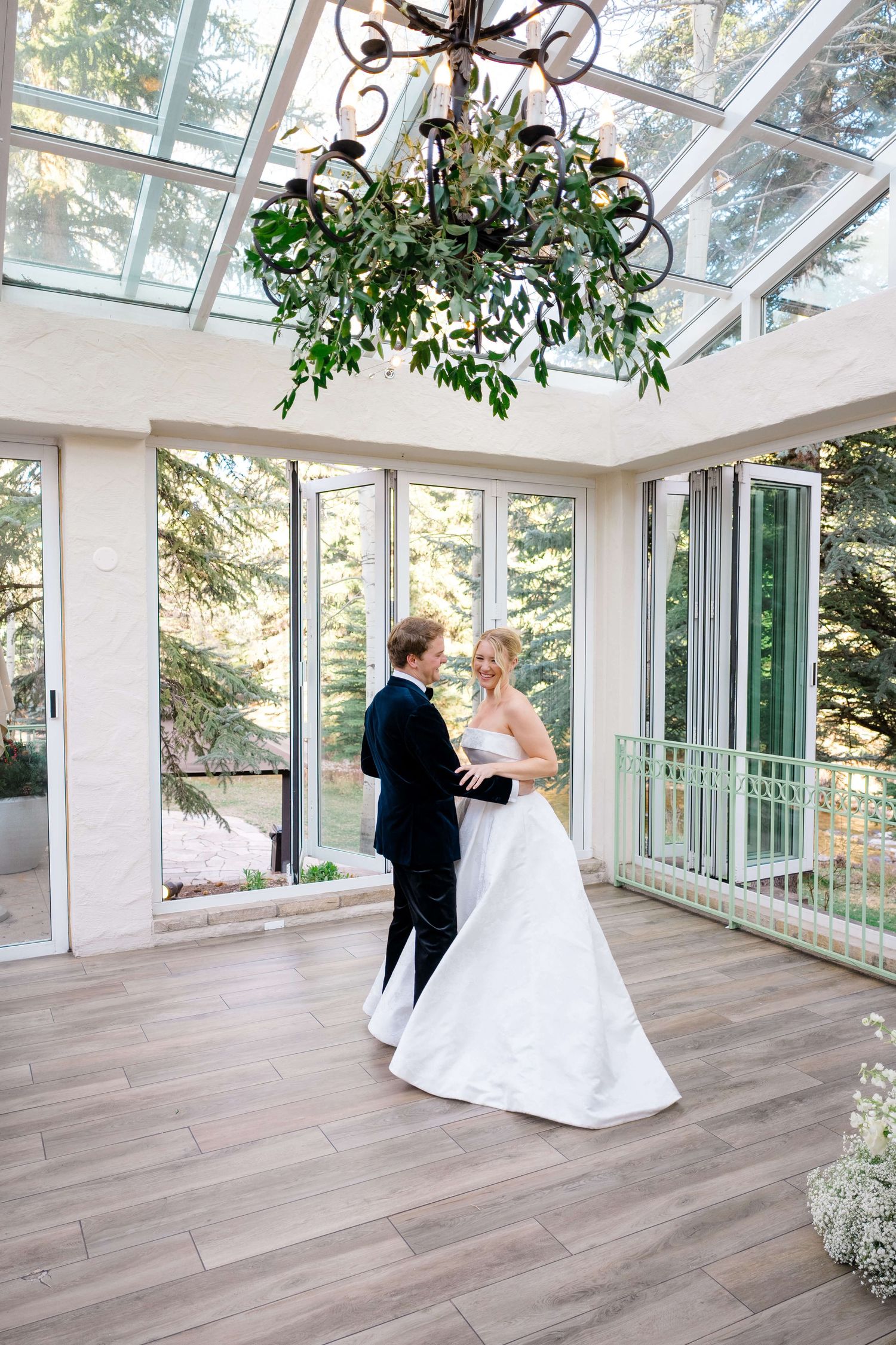 Bride and groom share their first dance in a sunlit glass conservatory with a floral chandelier.
