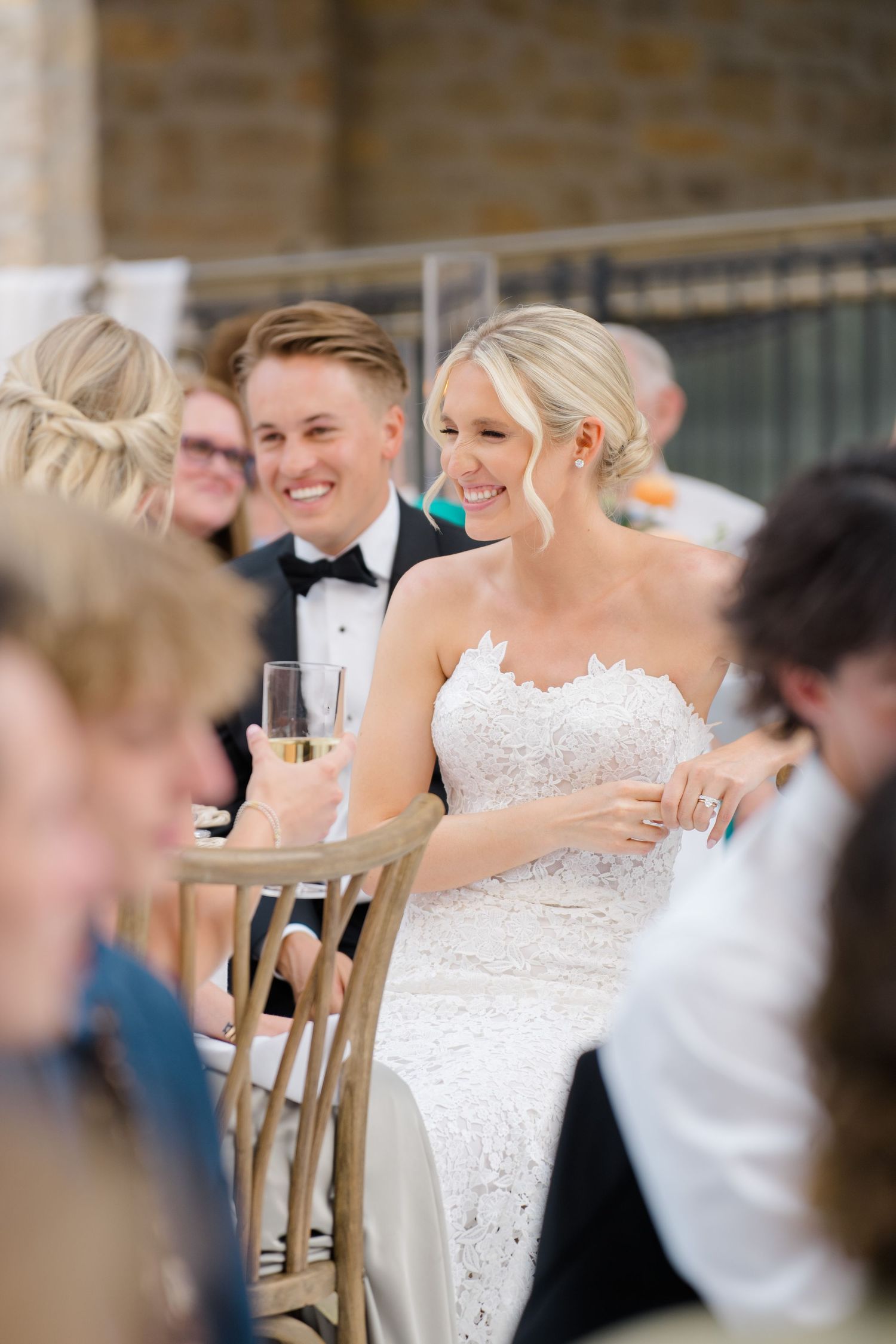 At Vail Arrabelle Wedding, Smiling bride in lace gown holding champagne seated beside groom in tuxedo at elegant wedding reception.