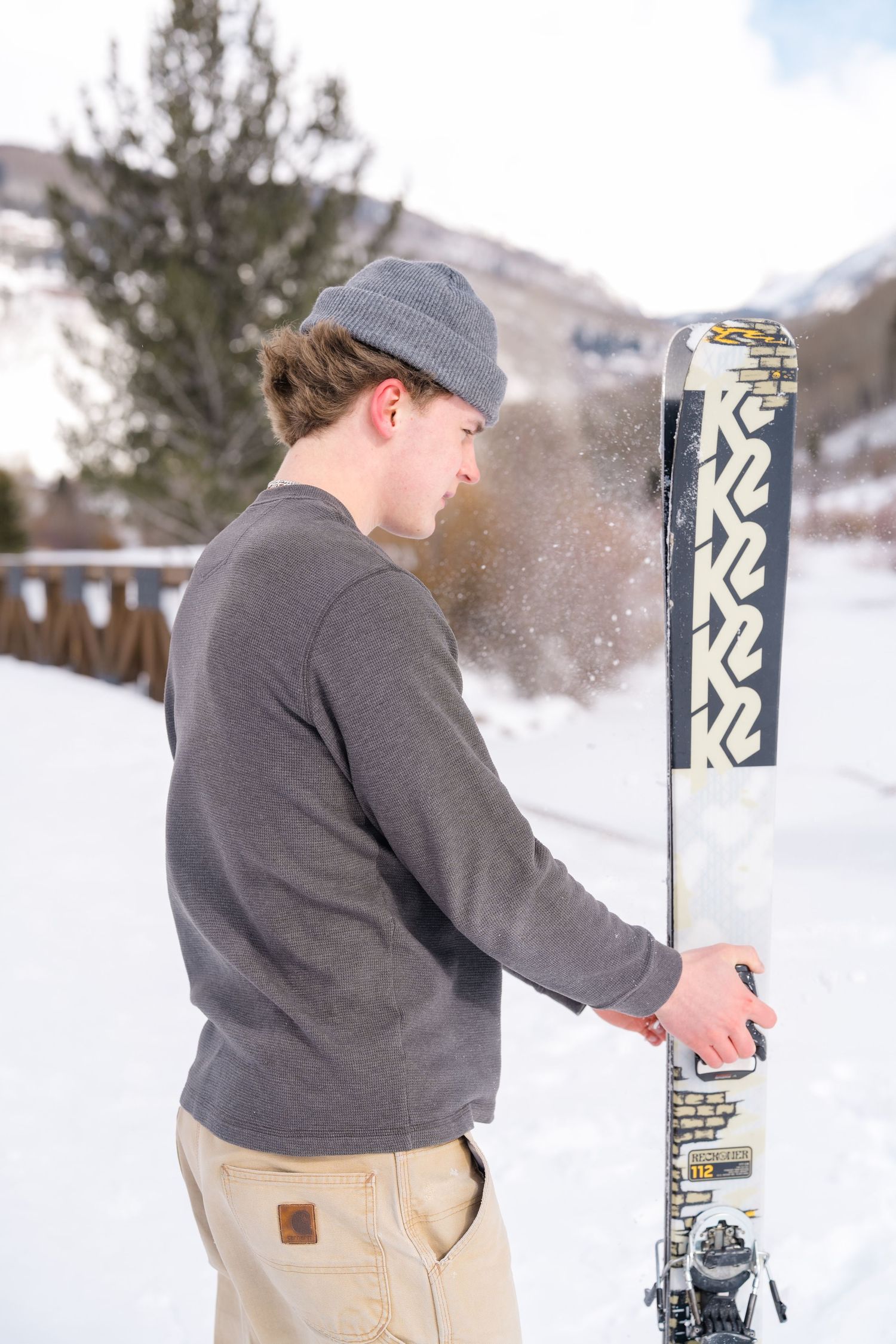 A person in a gray sweater and beanie brushes snow off their K2 ski on a winter mountain slope.