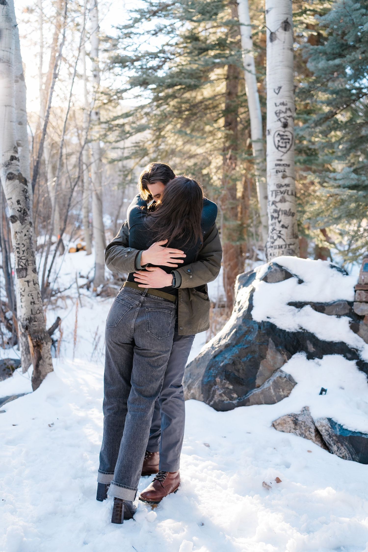 A couple embraces in a snowy forest setting with snow-covered trees and ground creating a romantic winter scene.