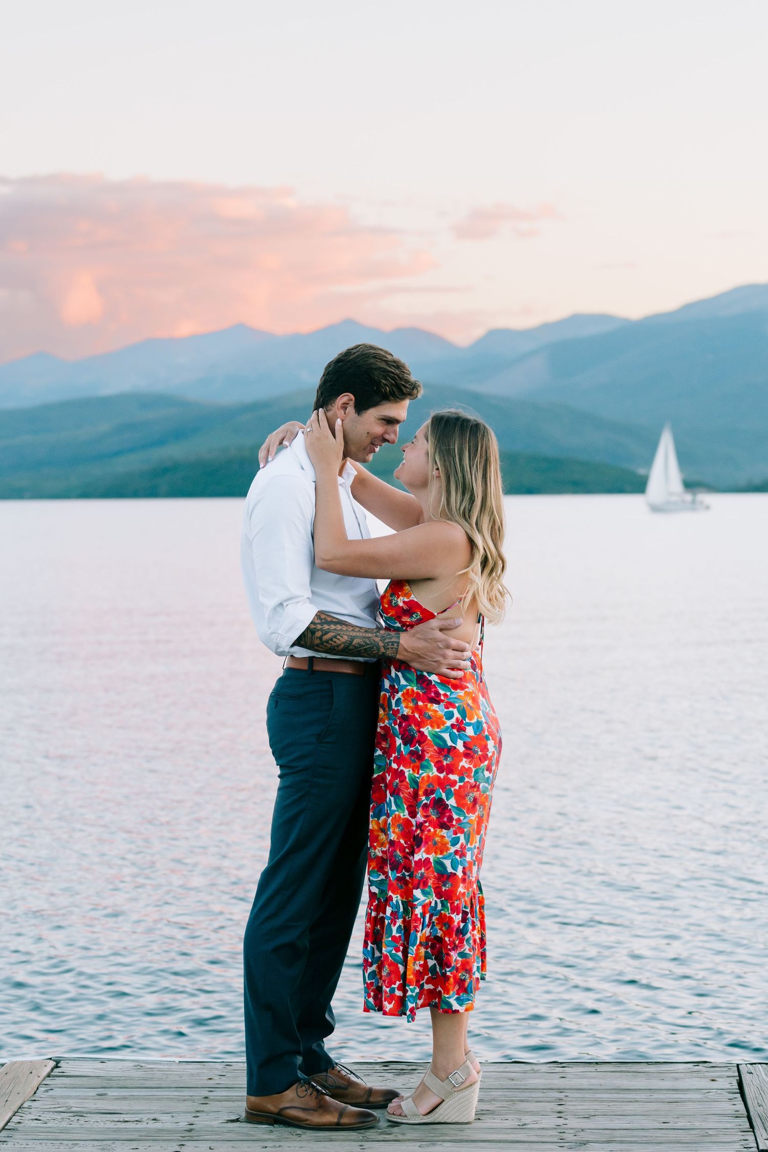 A romantic couple embraces on a wooden dock at sunset with mountains and a lake in the background.