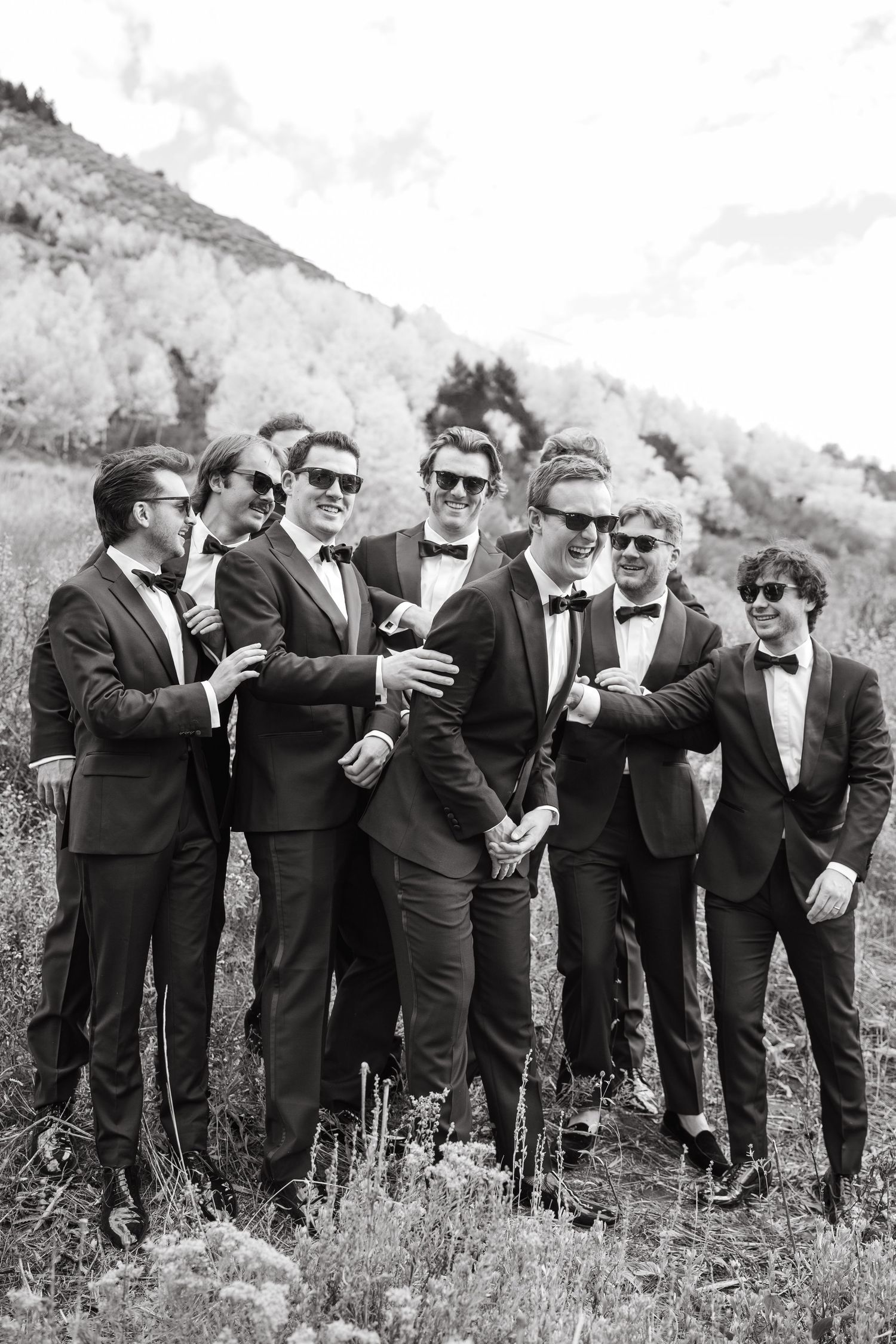 Groomsmen in black tuxedos and sunglasses pose together outdoors against a mountain backdrop.