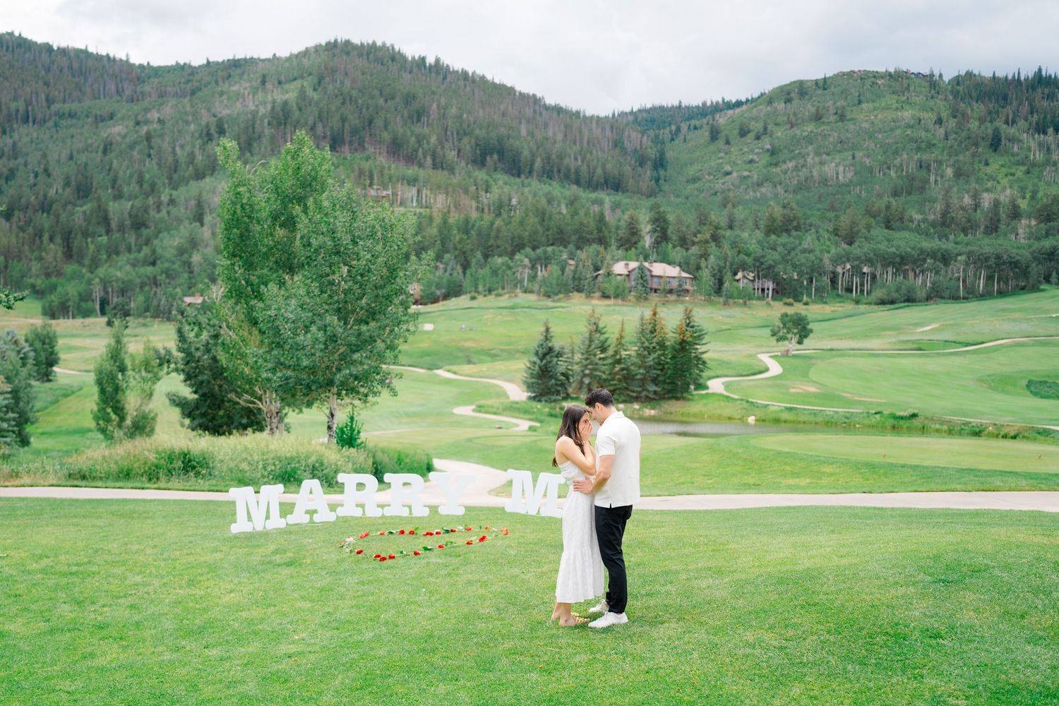 A couple embraces on a scenic golf course with rolling green hills and mountains in the background.