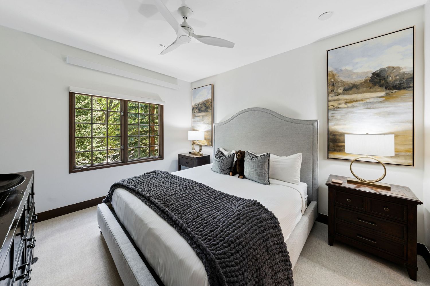 Modern bedroom with gray upholstered bed, dark wood nightstands, ceiling fan, and large window overlooking lush greenery.