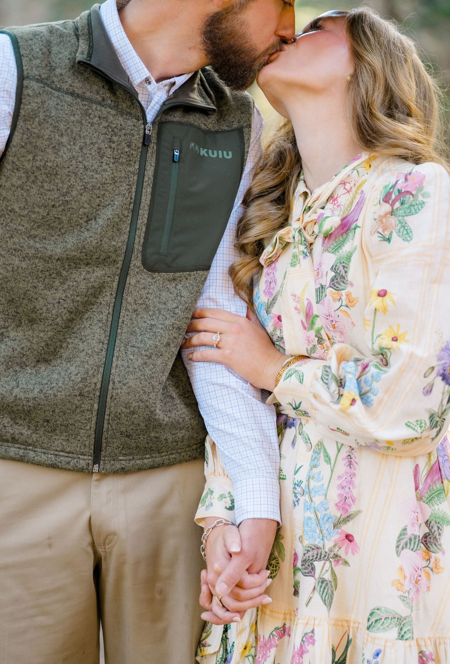 Couple sharing a romantic kiss, woman in floral dress holding hands with man in green KUIU vest outdoors.