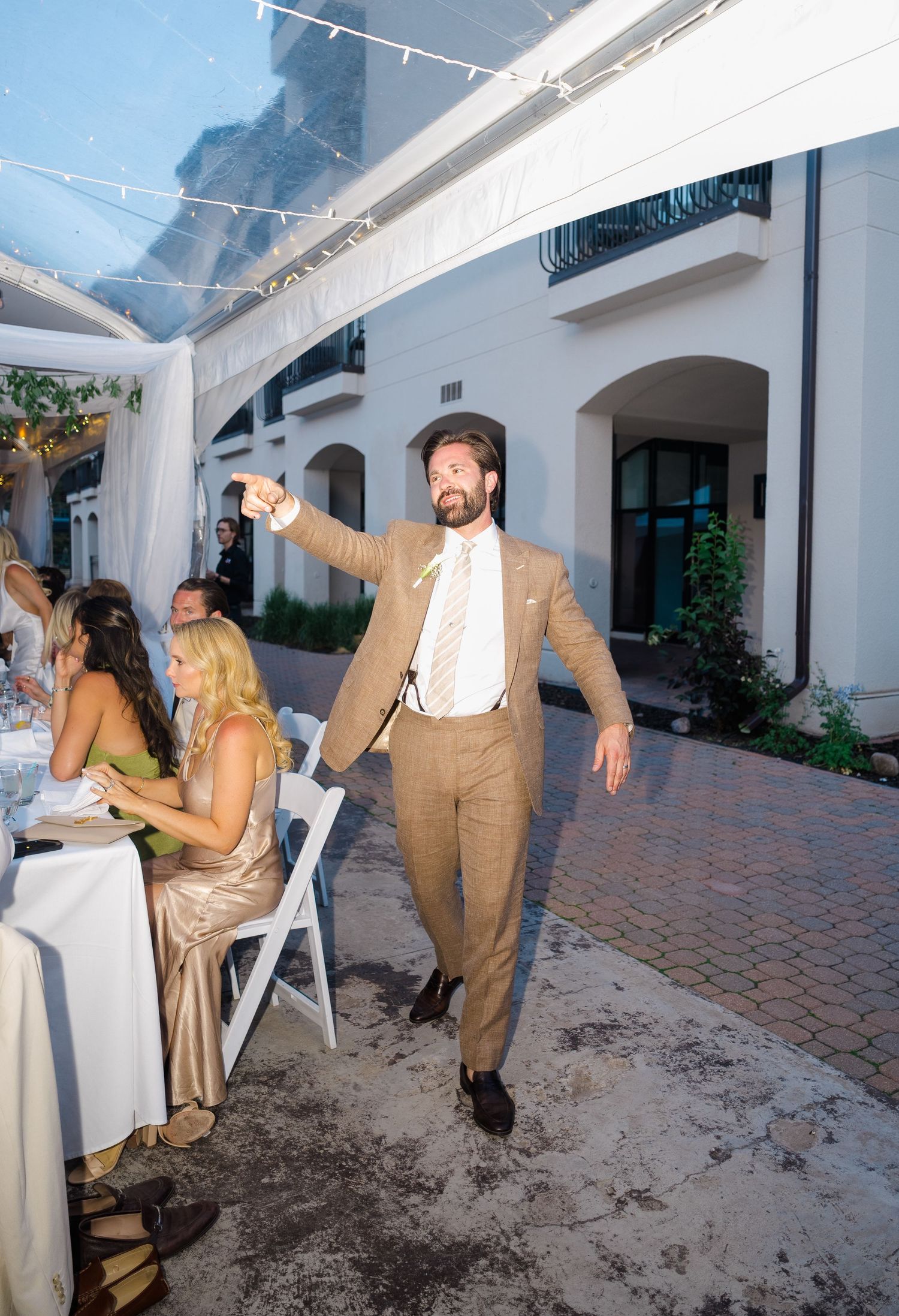 Man in tan suit pointing at outdoor wedding reception dinner at elegant white venue.