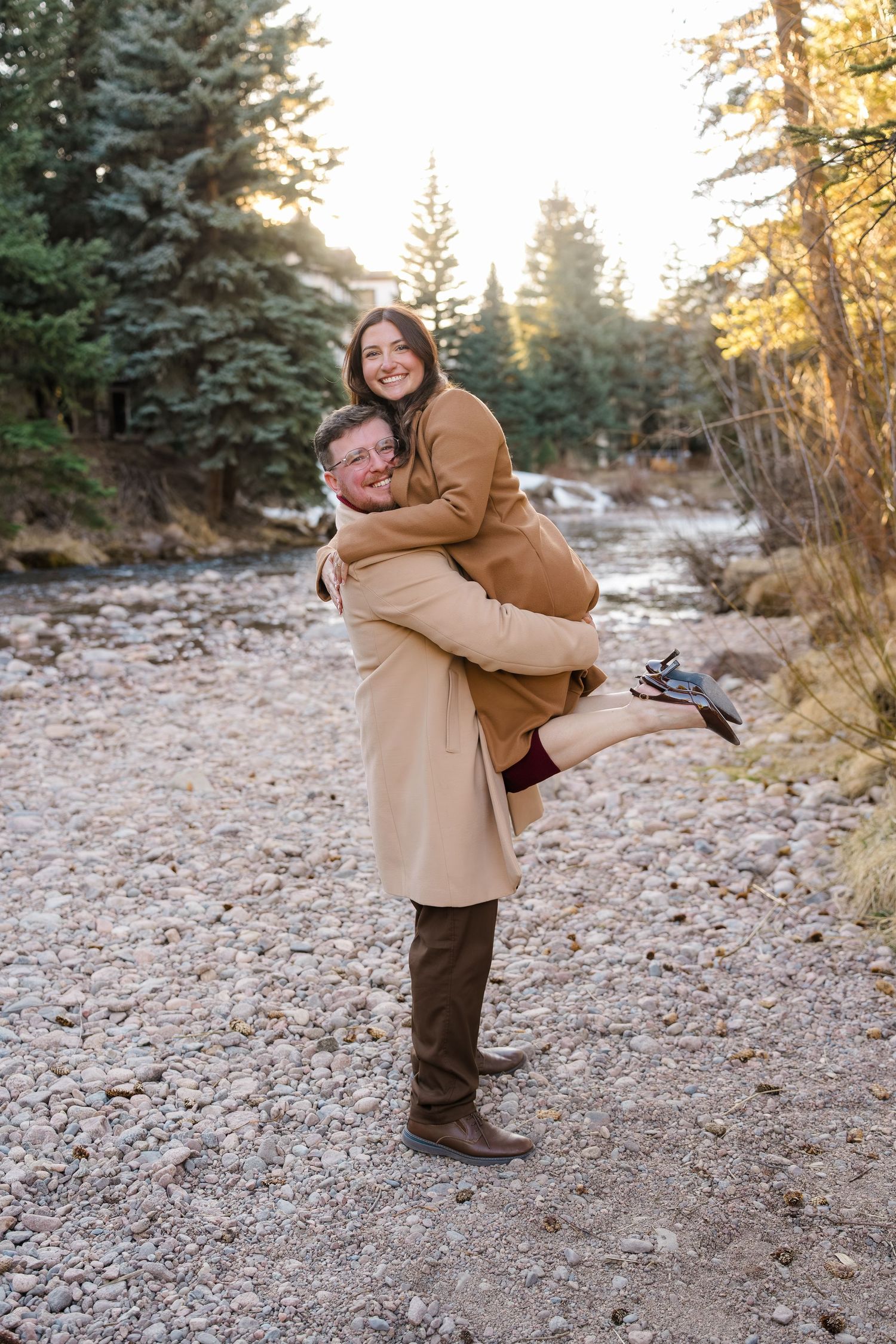 A joyful couple embraces outdoors, man lifting woman near a rocky riverbank surrounded by pine trees in warm autumn light.