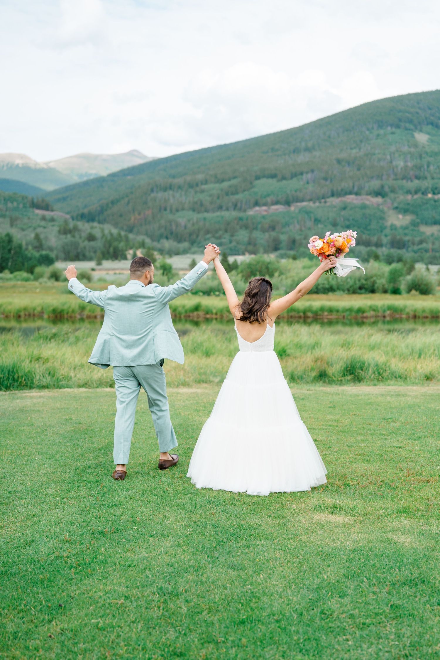 A couple in wedding attire dances playfully in a green meadow with mountains in the background.