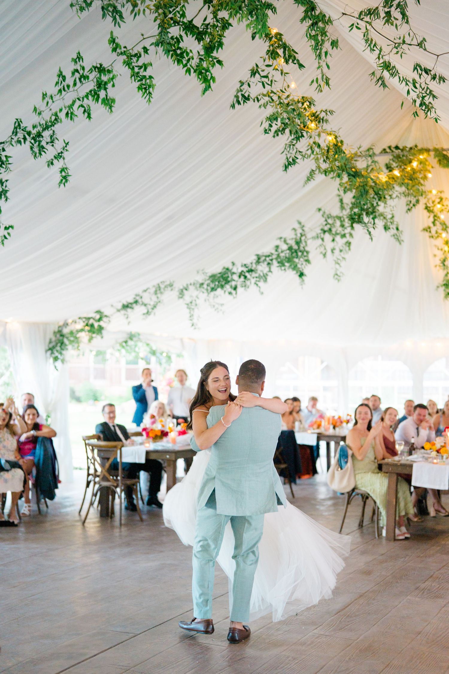 Wedding reception dance under a white tent decorated with hanging greenery and twinkling lights.