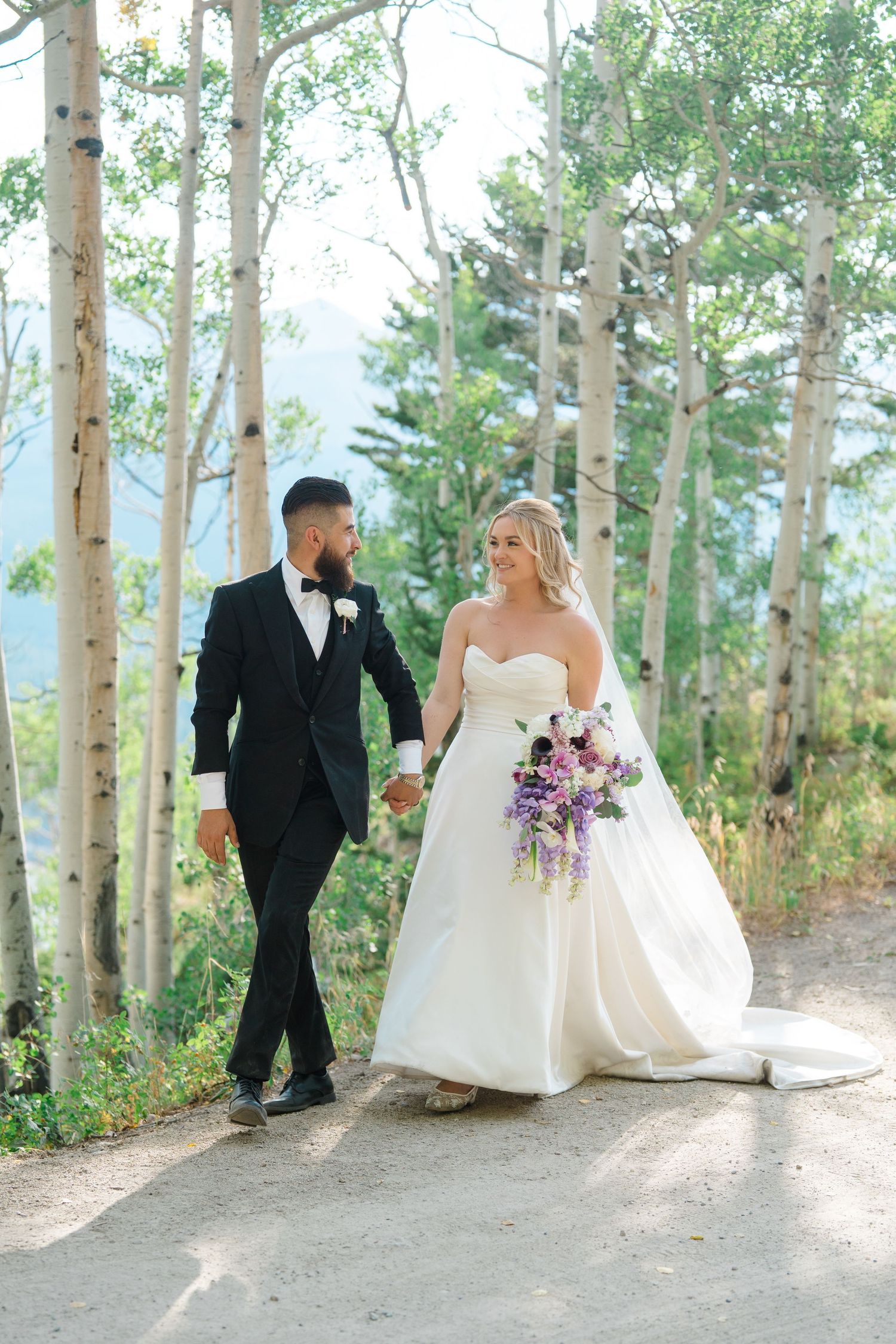A bride in white gown holds purple bouquet while walking with groom through aspen trees on mountain path.