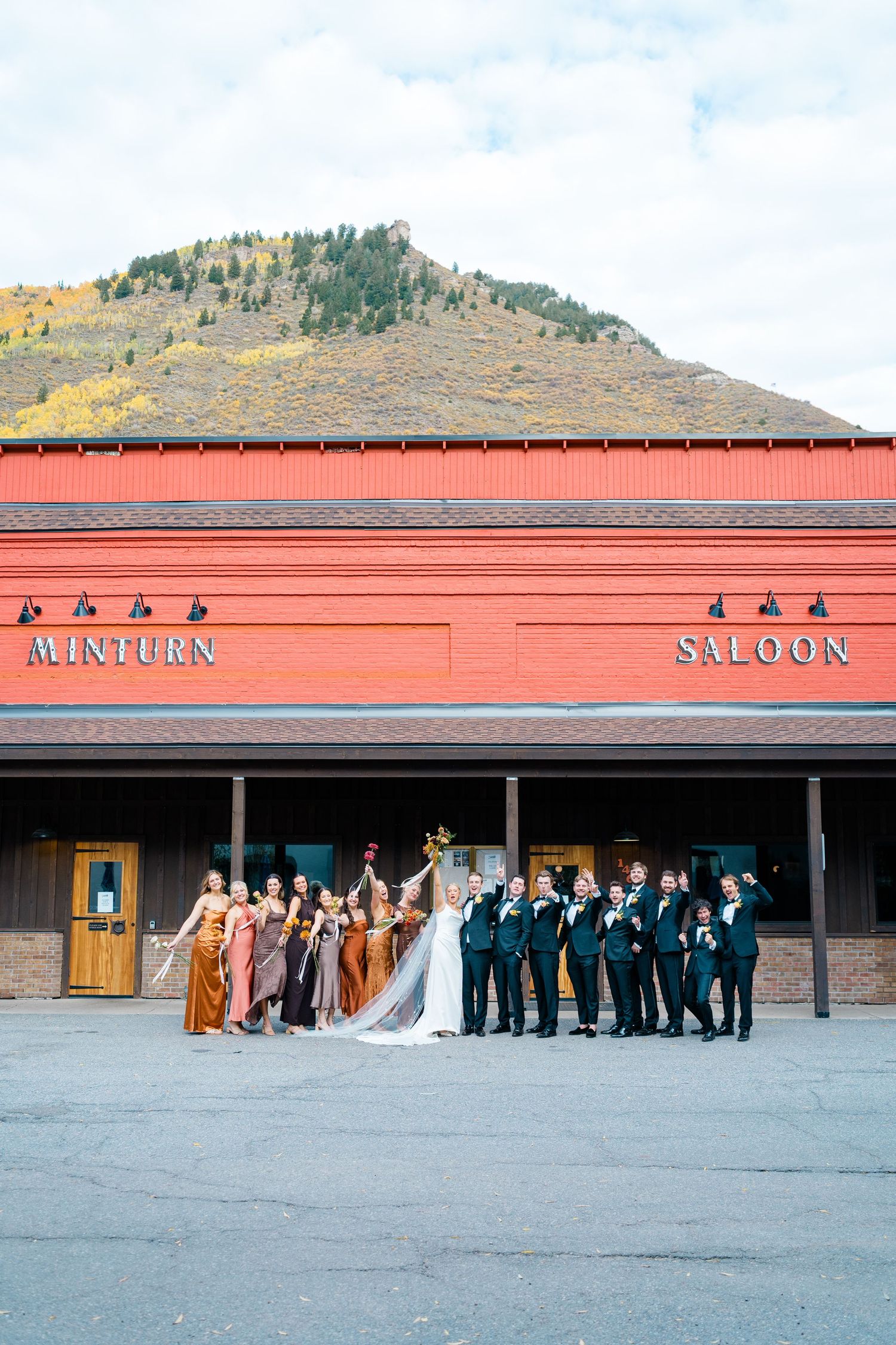 Wedding party poses for group photo outside Mine Link Saloon with mountain backdrop.